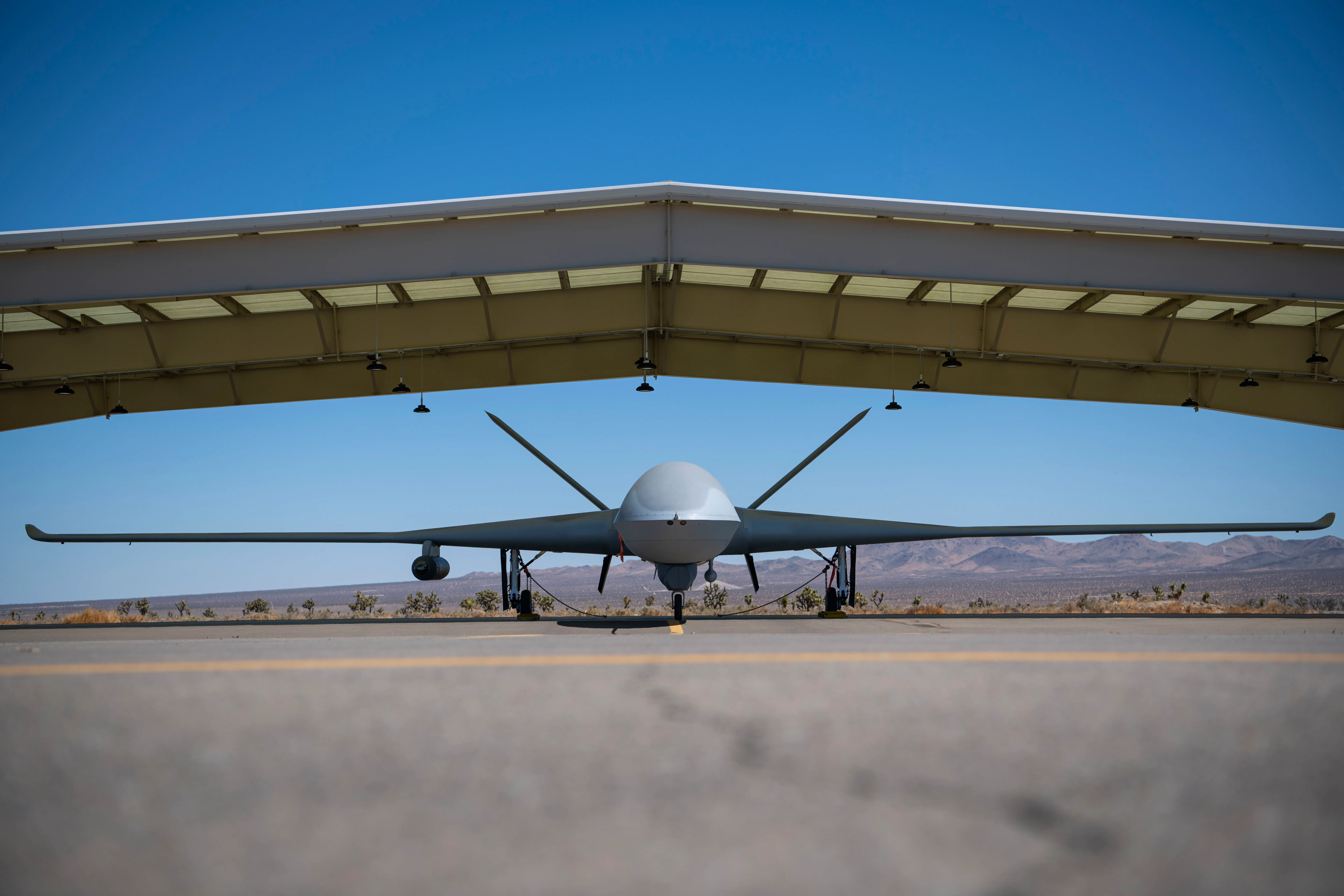 An MQ-20 Avenger uncrewed aircraft is seen parked under an awning in the desert.