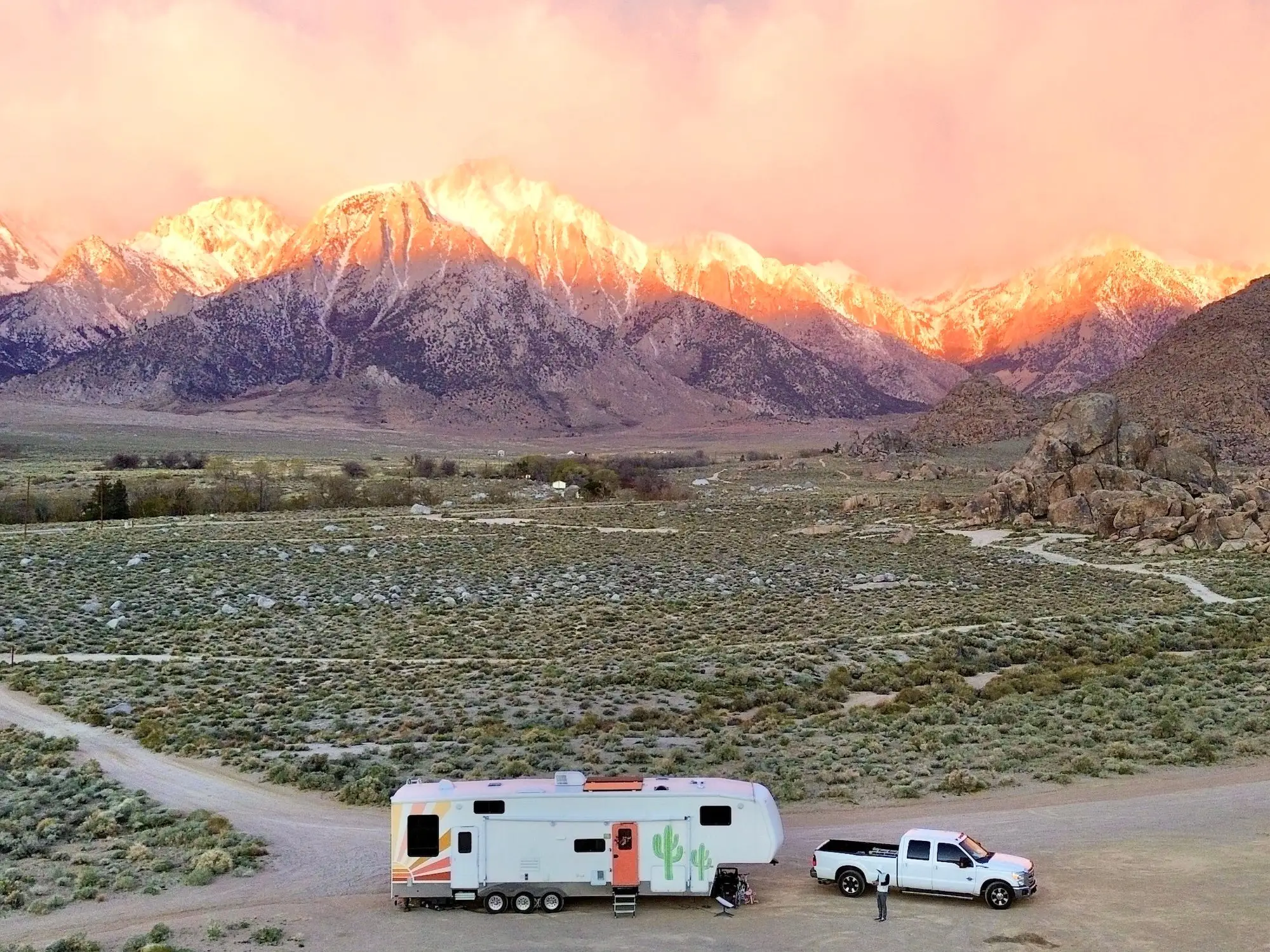 A camper trailer and white pickup sit on a desert road below sunlit snowy mountains and pink clouds.