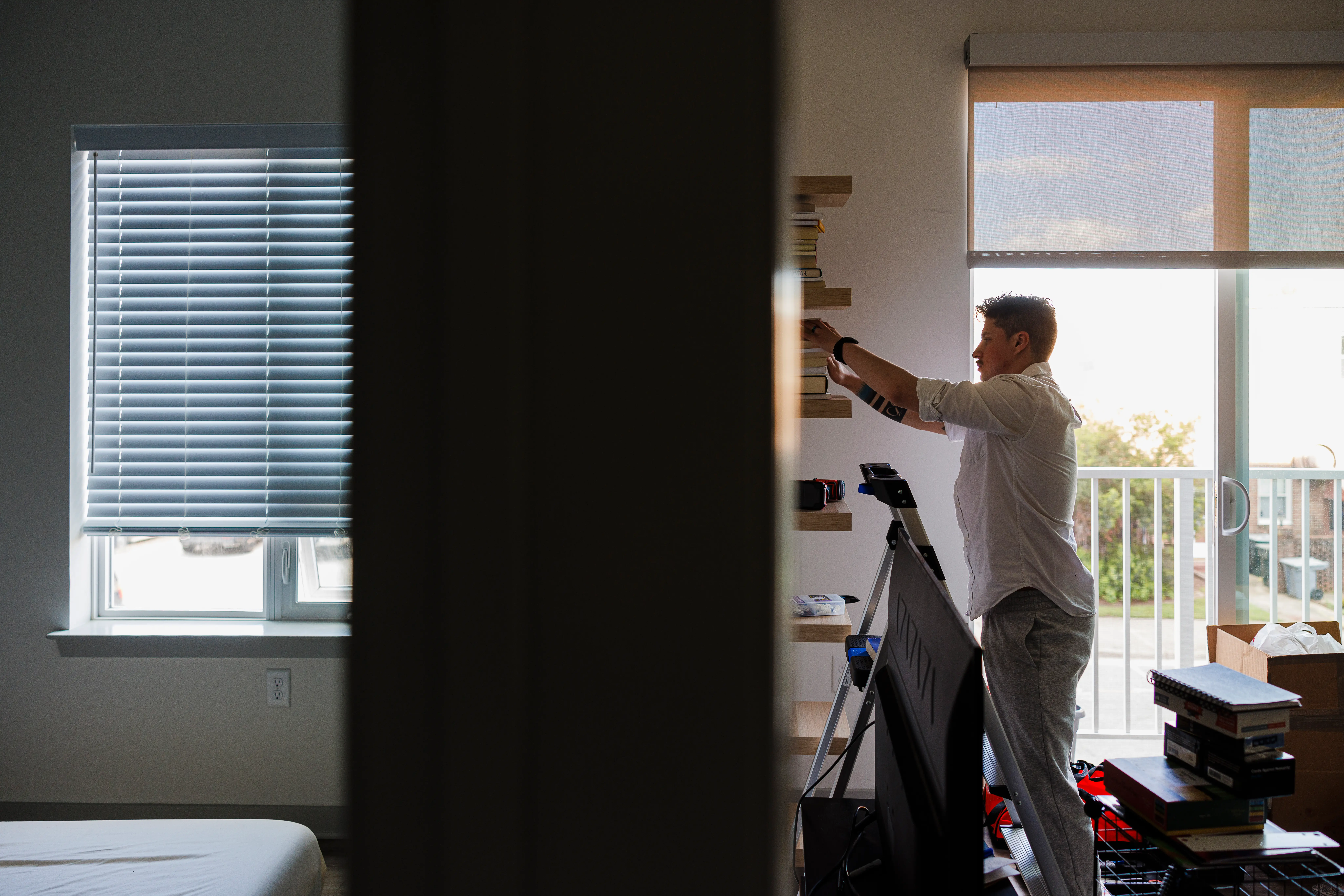 Person organizes items on wall shelves in a sunlit apartment room beside a window and tripod.