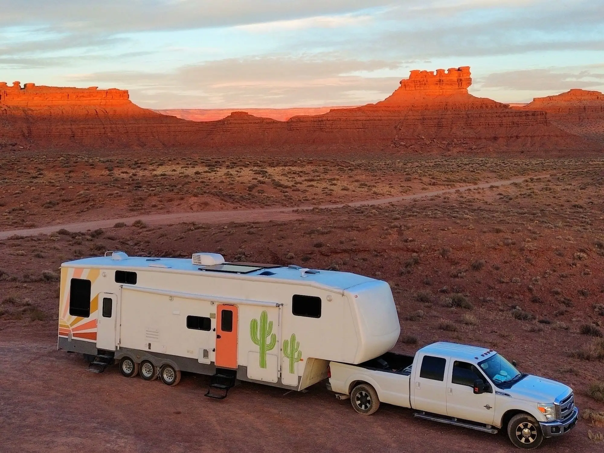 White pickup truck towing a fifth-wheel RV parked in a red rock desert landscape under a blue sky.