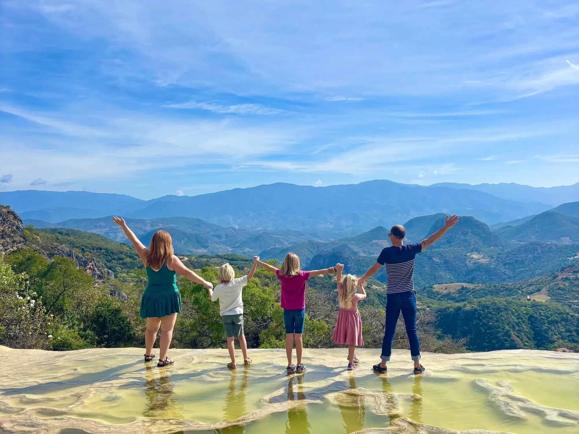 Five people hold hands on travertine mineral pools overlooking a broad mountain valley under a blue sky.