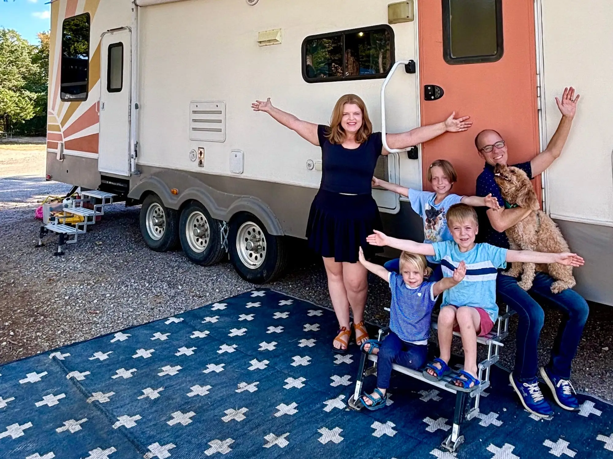 Five people and a dog pose with raised arms outside a large travel trailer on a patterned outdoor rug.