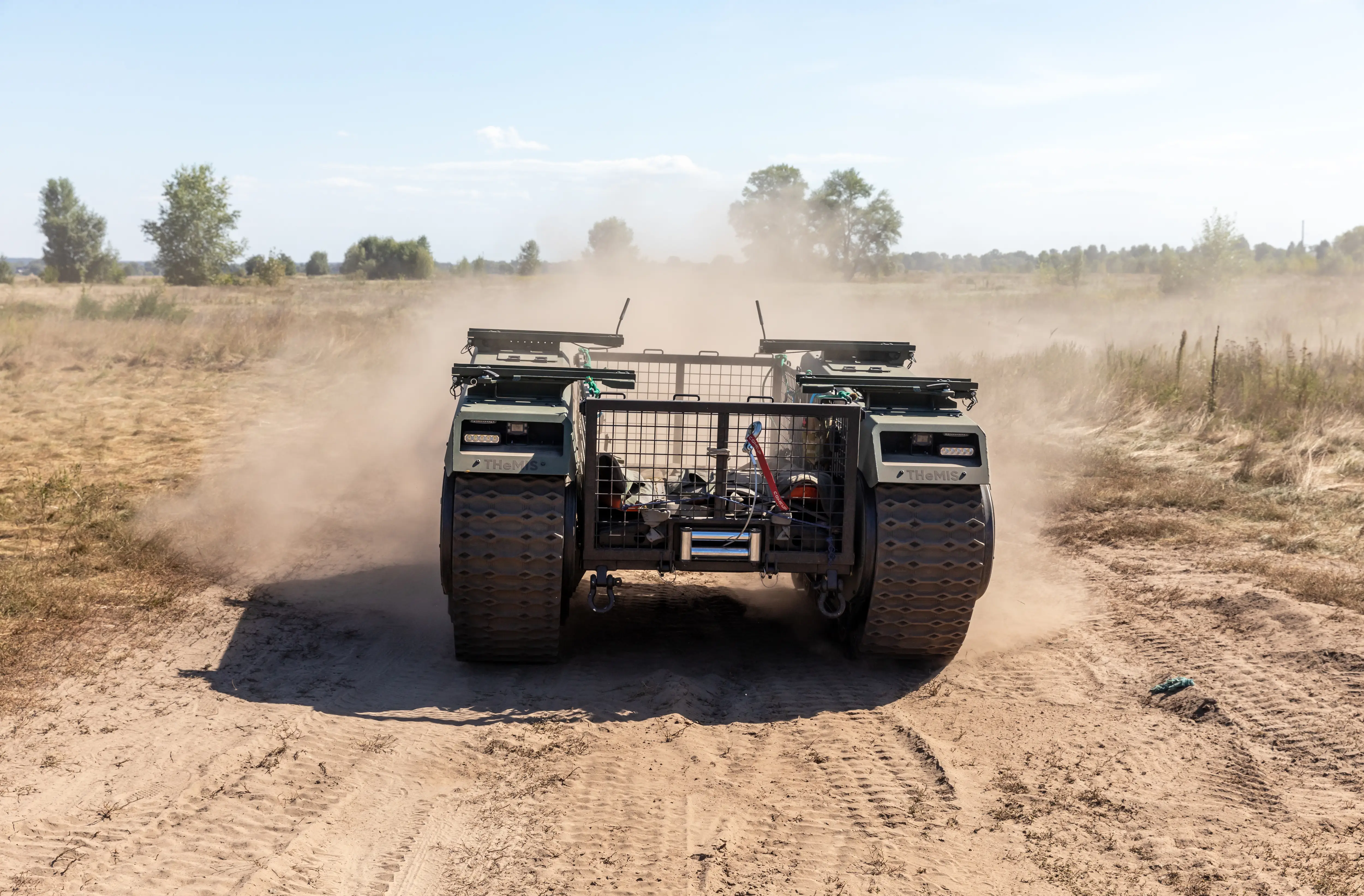 Tracked robotic vehicle drives along a dusty rural dirt road with fields and trees in the background.