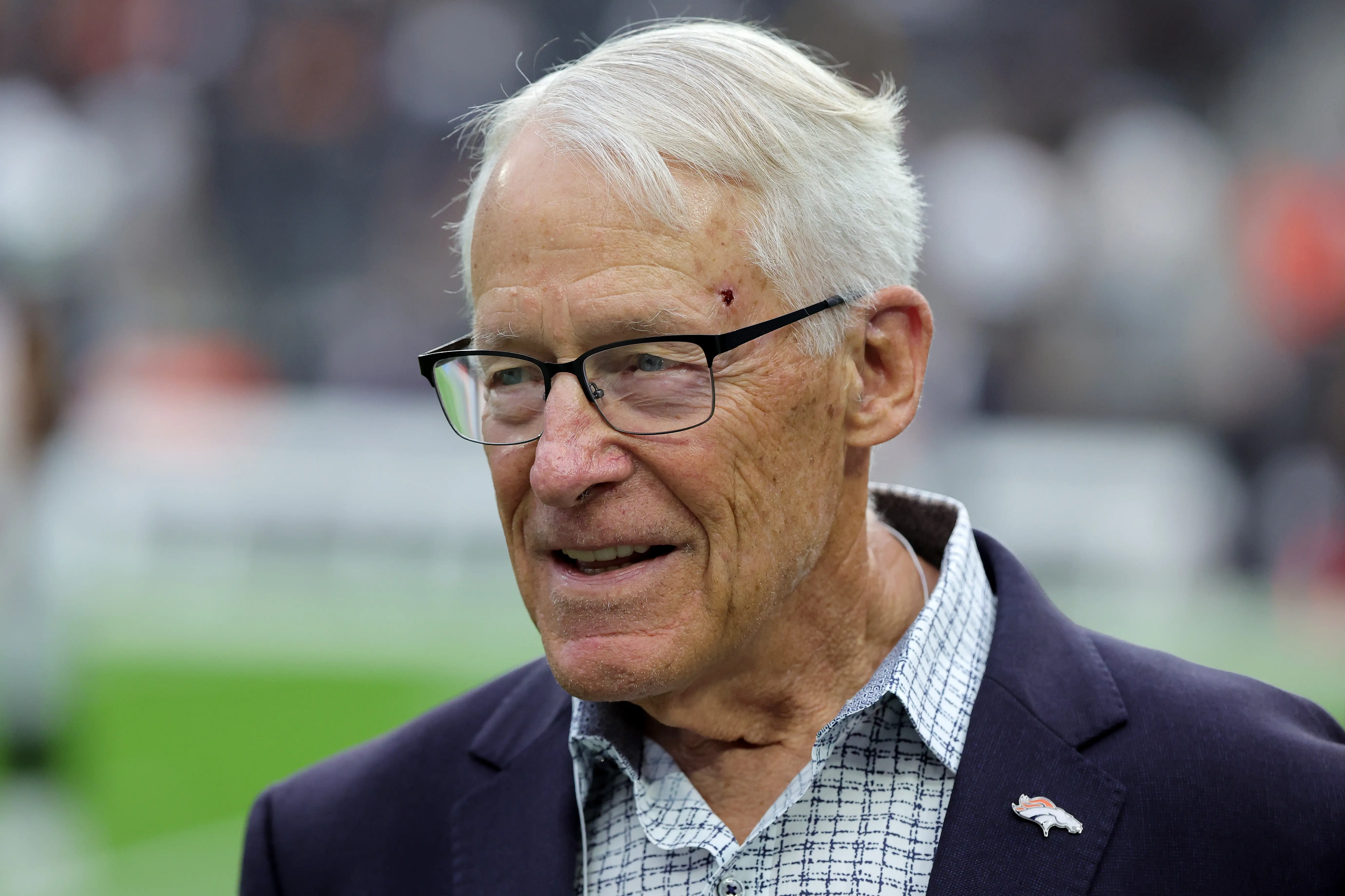 White-haired person wearing glasses and a blazer stands on a football field with a Denver Broncos lapel pin.