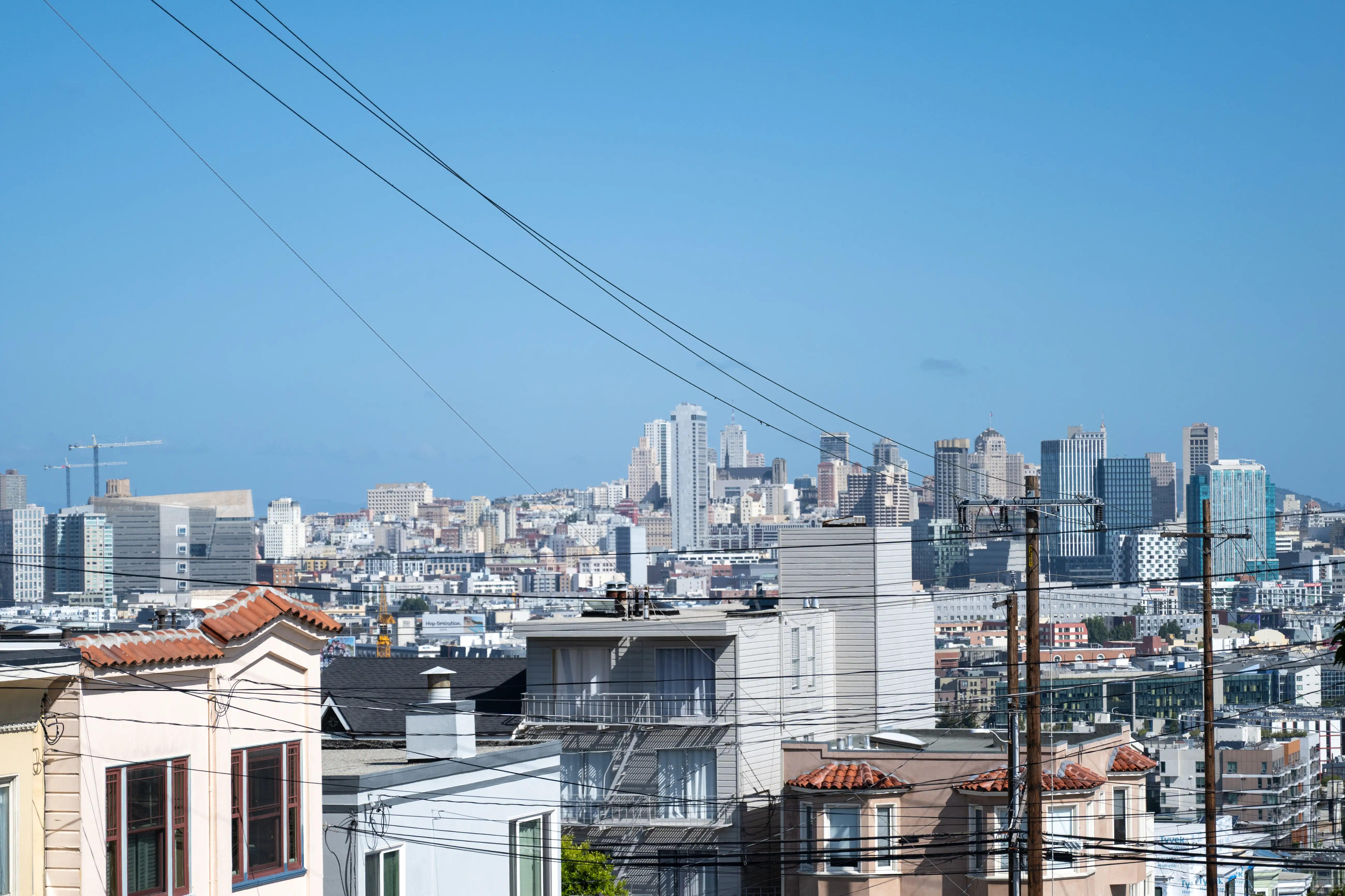 Urban skyline and residential rooftops with utility poles and overhead wires under a clear blue sky.