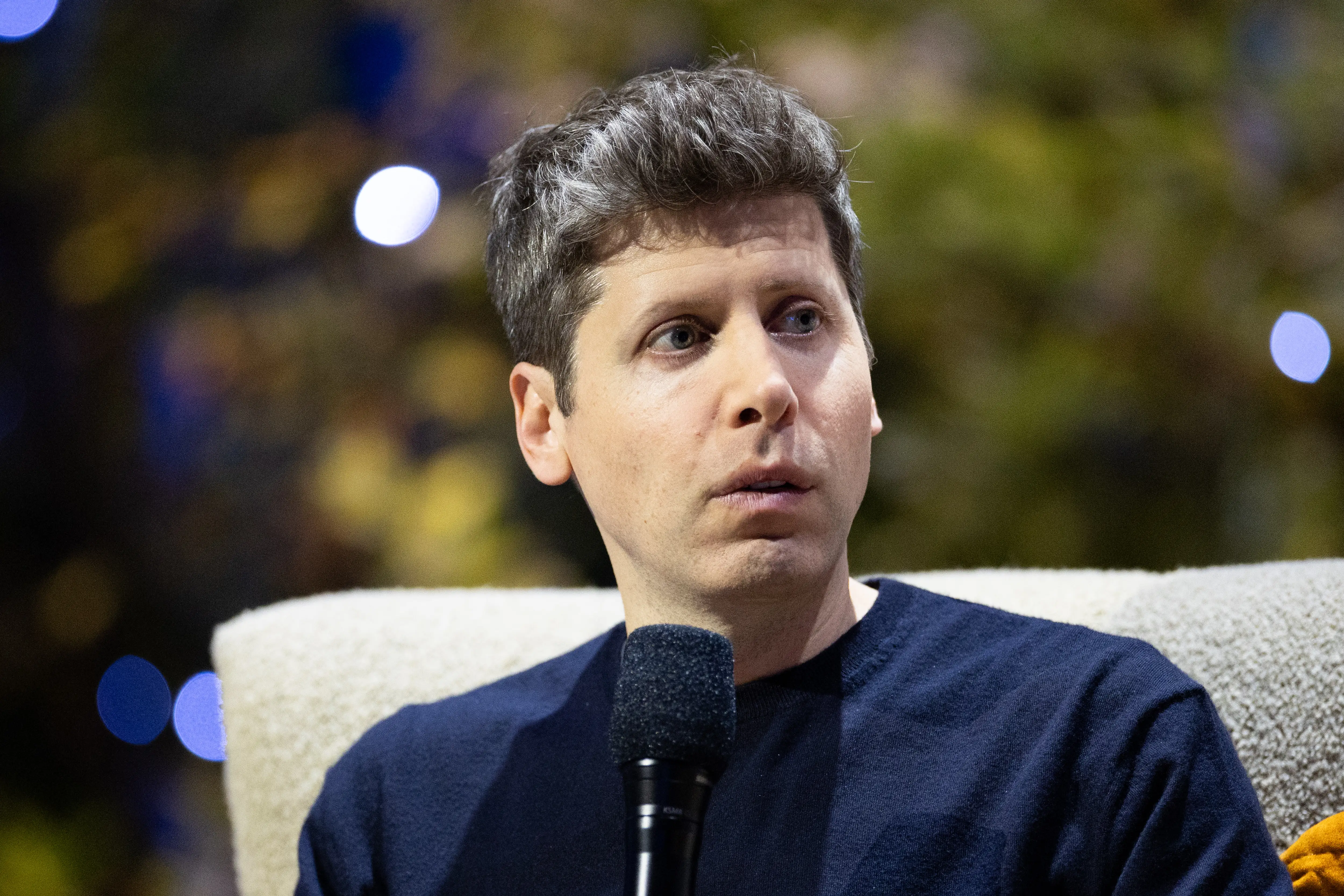 Sam Altman holds a microphone while seated on a light-colored chair during an indoor speaking event.