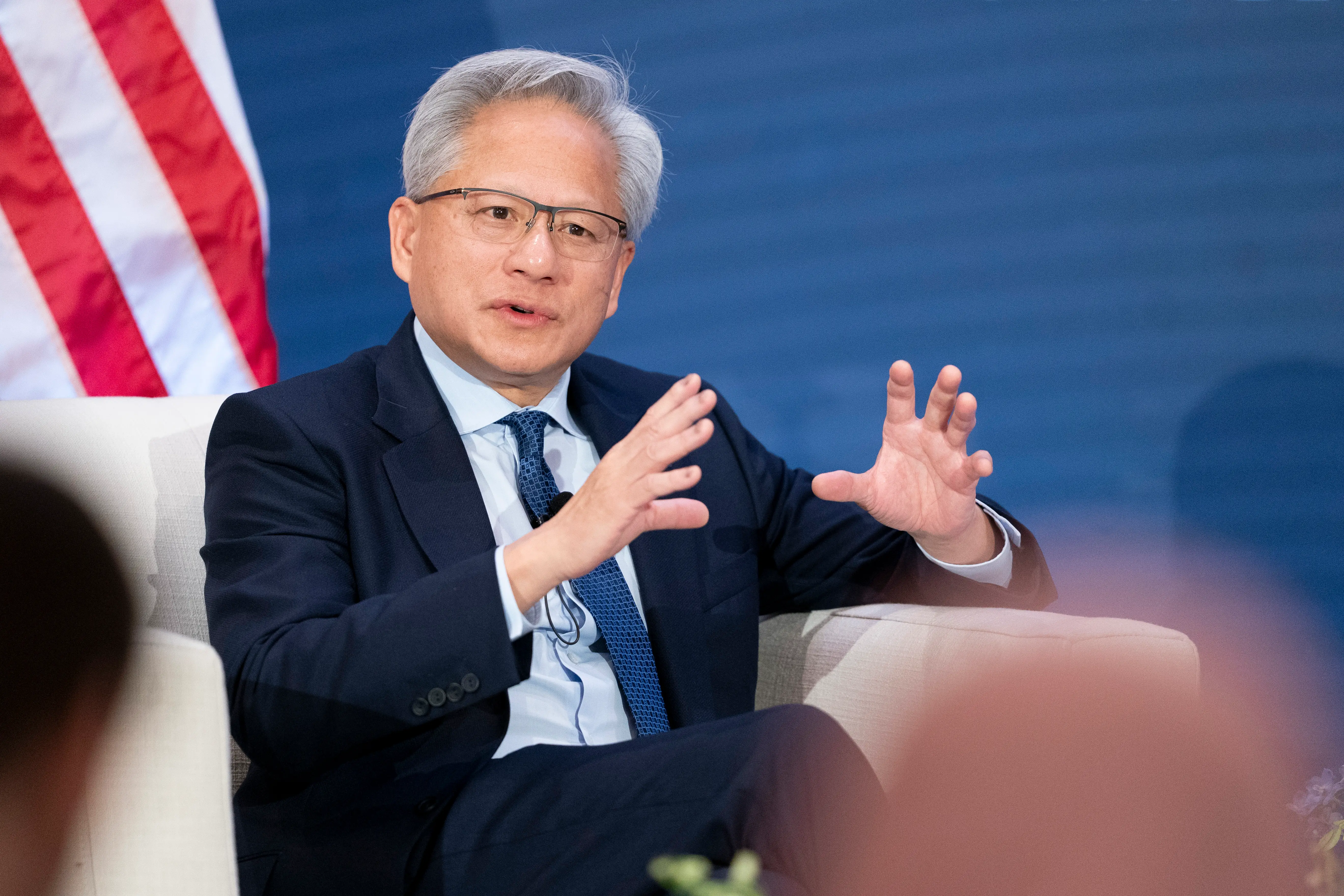 Jensen Huang sits onstage in a suit, gesturing while speaking beside a visible United States flag.