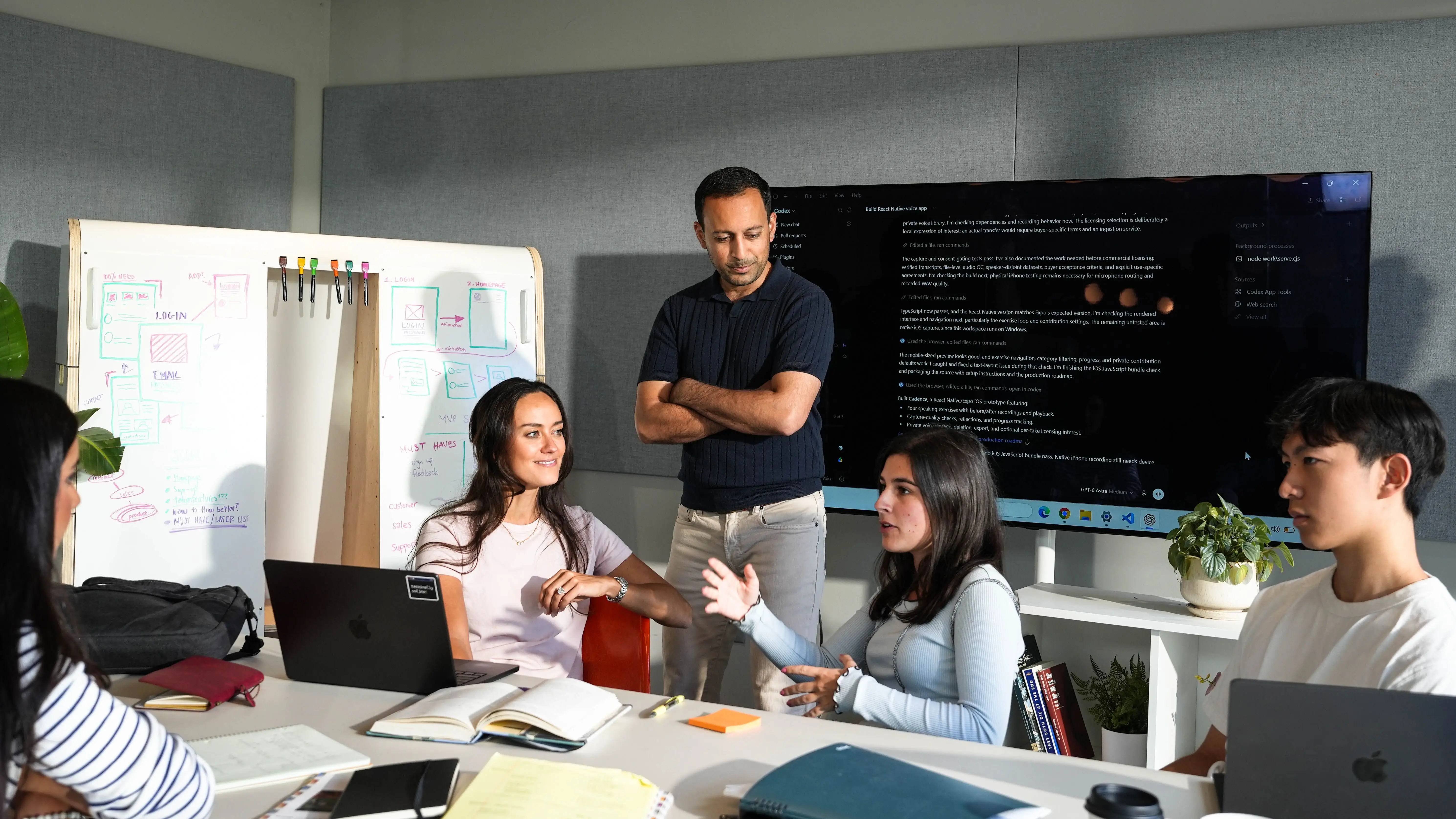 Group meeting in a conference room with laptops, notebooks, a whiteboard, and a large display showing code.