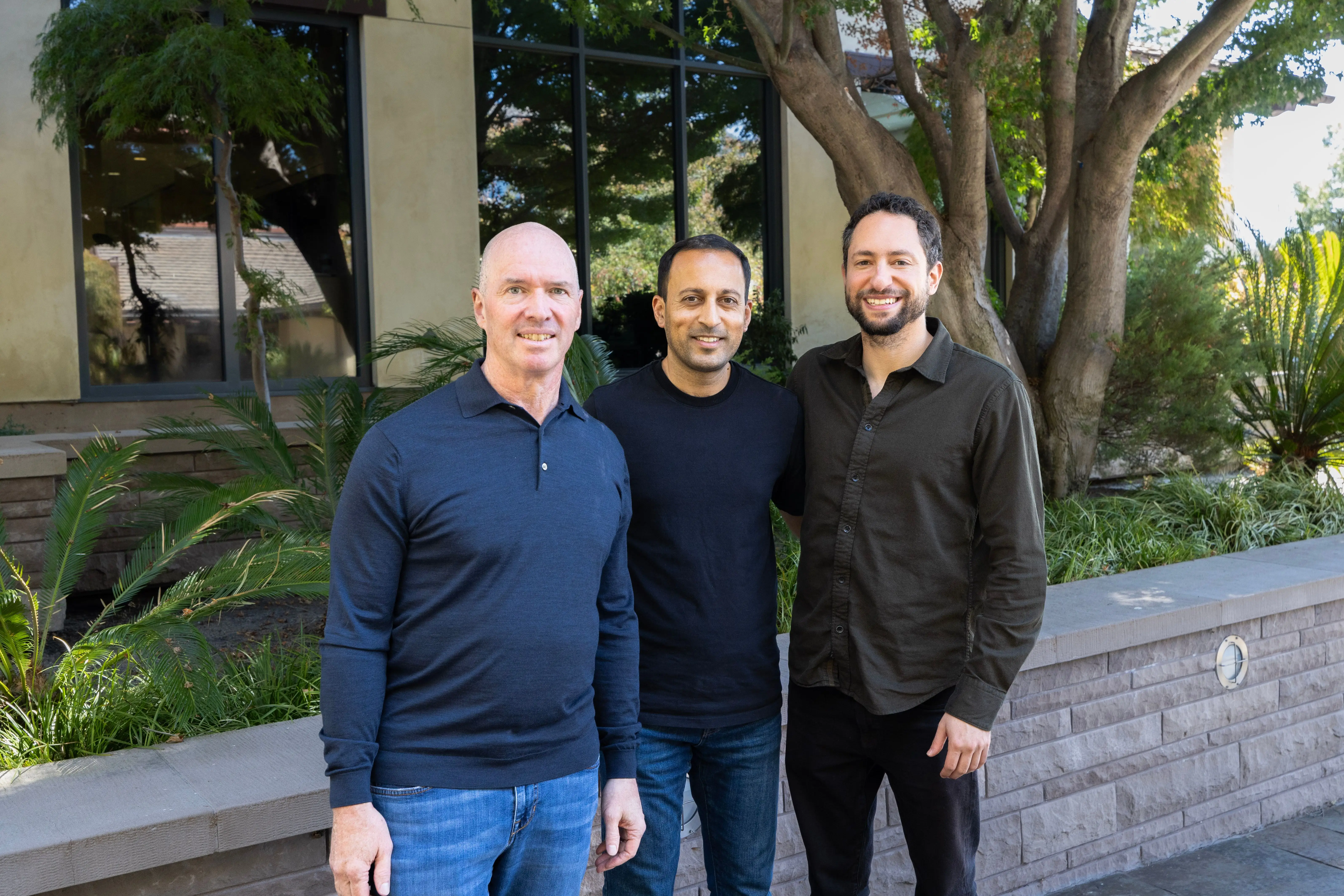 Three people stand together outside a landscaped building with large windows and trees.