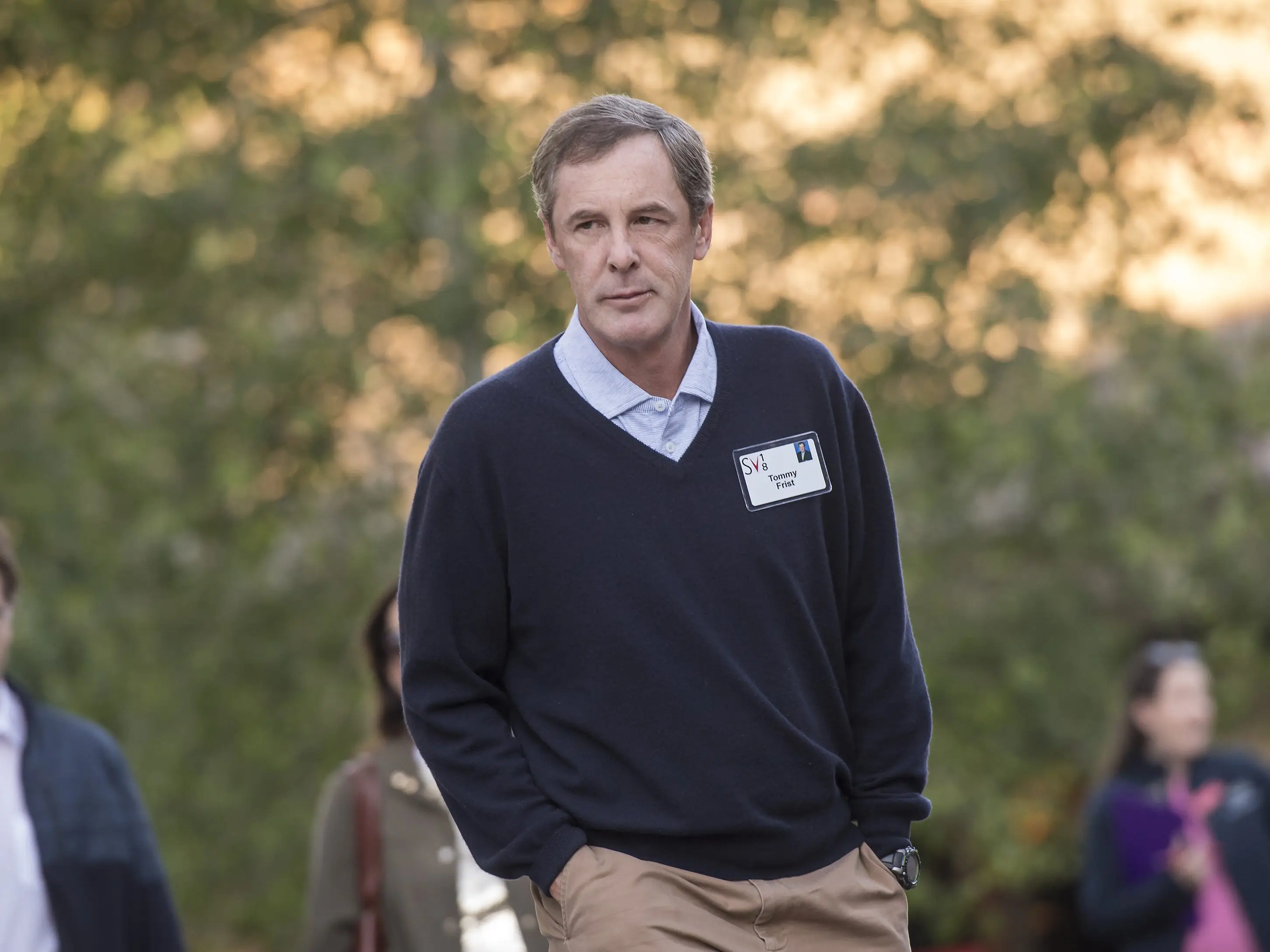 Person wearing a name badge stands outdoors at a blurred gathering with other attendees in the background.