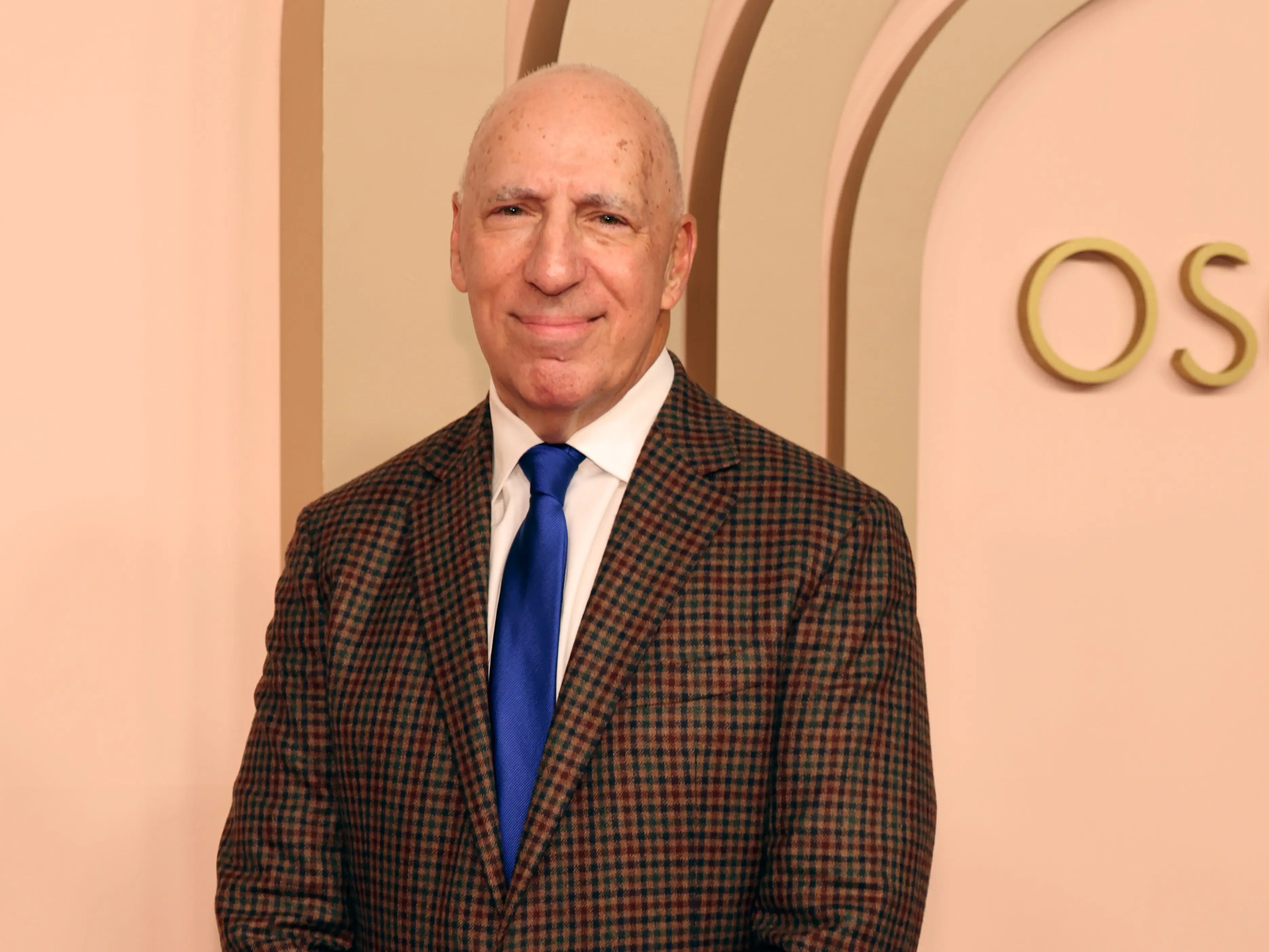 Steven Rales in a checked blazer and blue tie poses against a peach event backdrop.