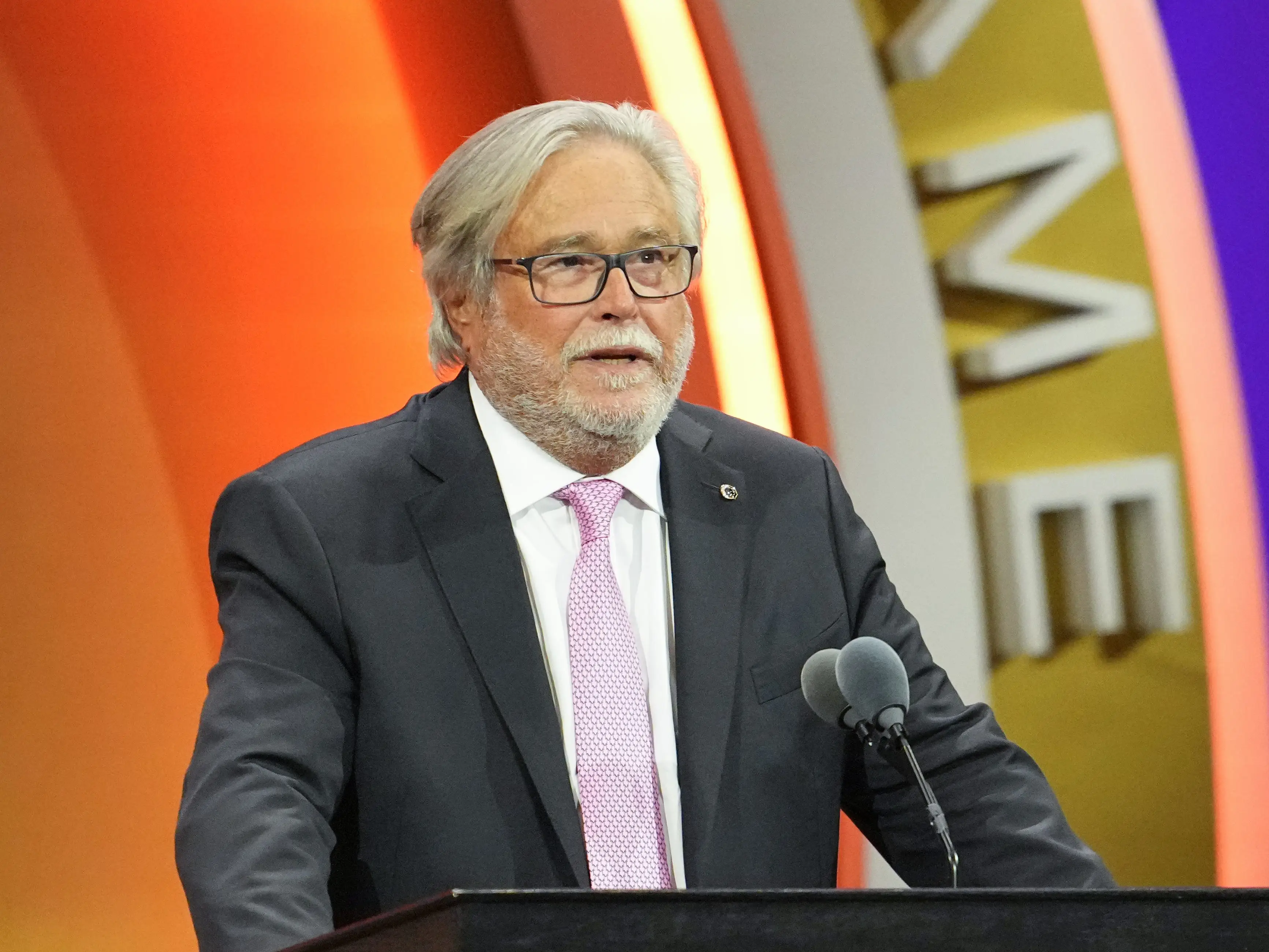 Micky Arison in a suit stands at a podium on a stage with a Miami Heat logo behind him.