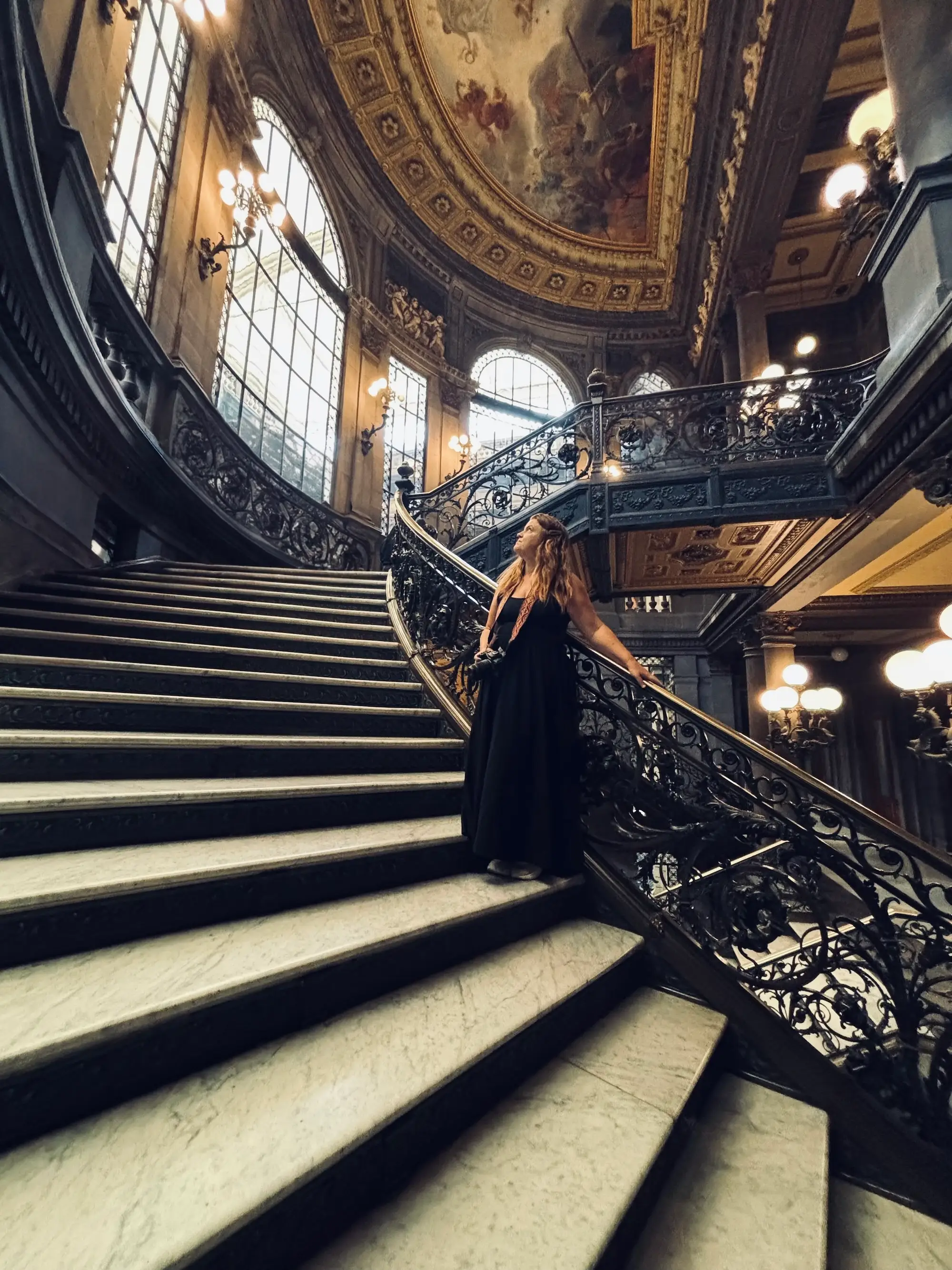 A person in a long black dress stands on an ornate marble staircase inside a grand historic interior.
