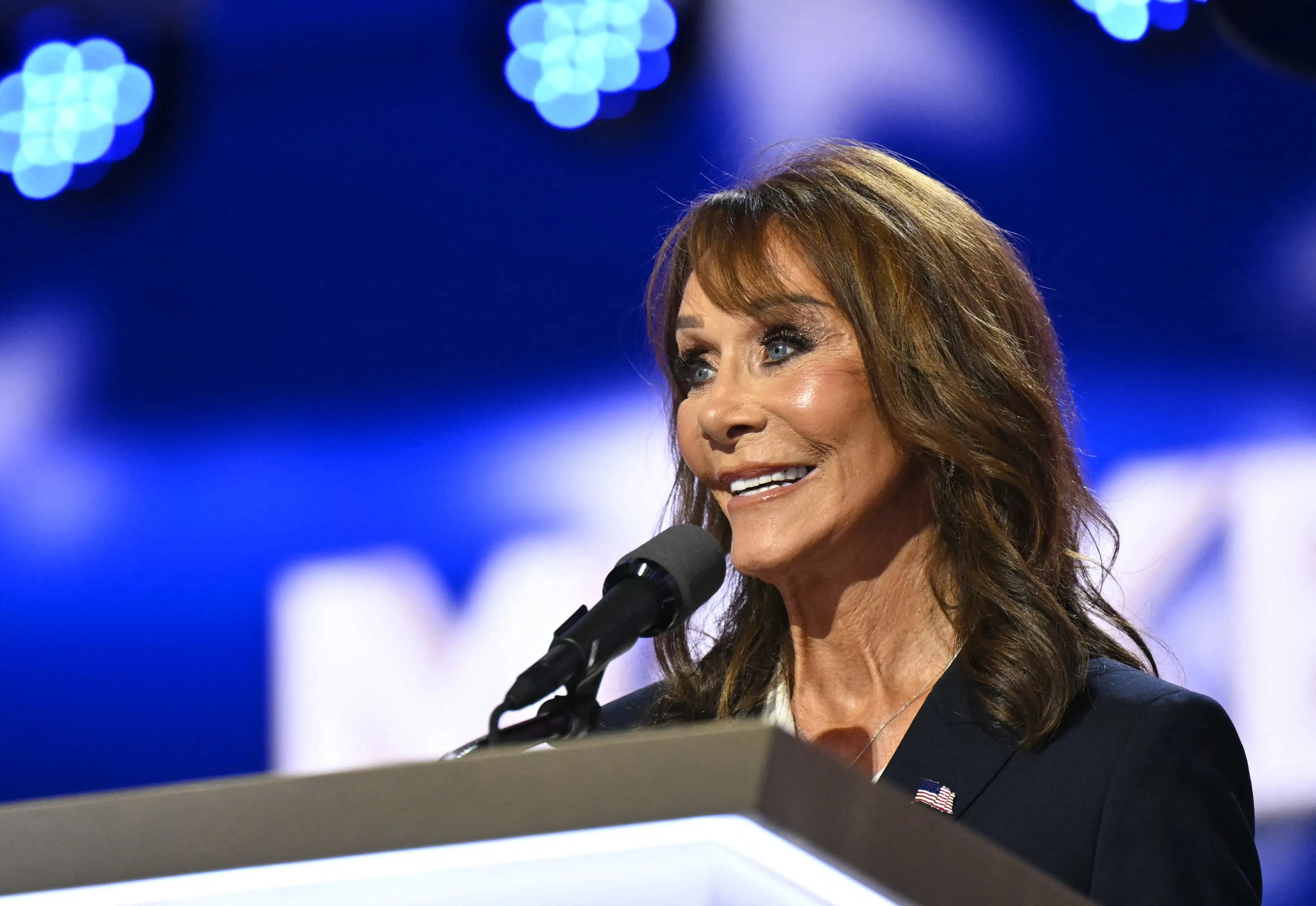 Speaker with an American flag lapel pin addresses a microphone at a blue-lit podium.
