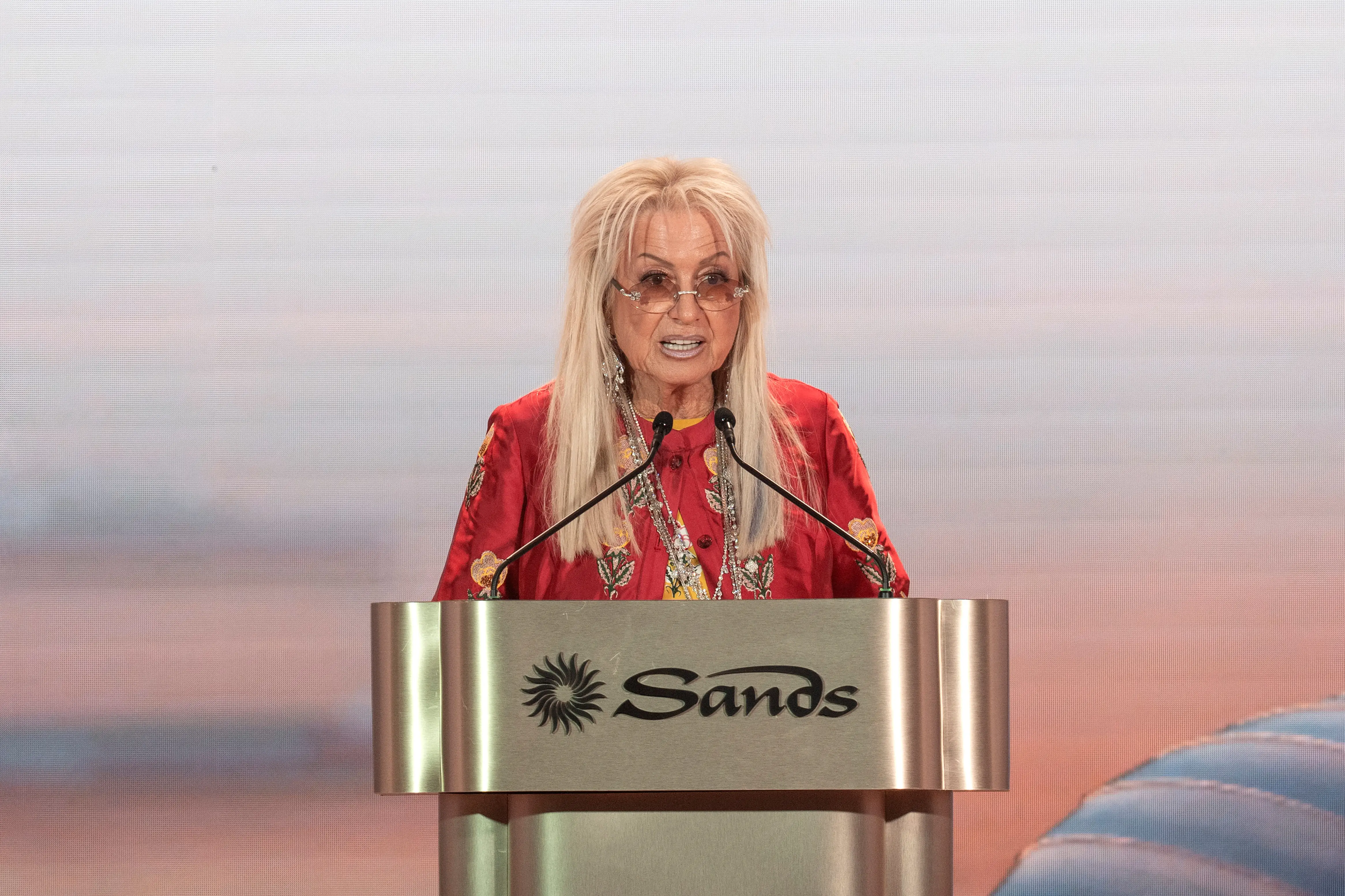 Miriam Adelson, in red attire, addresses the microphones at a podium displaying the Sands logo against a large-screen backdrop.