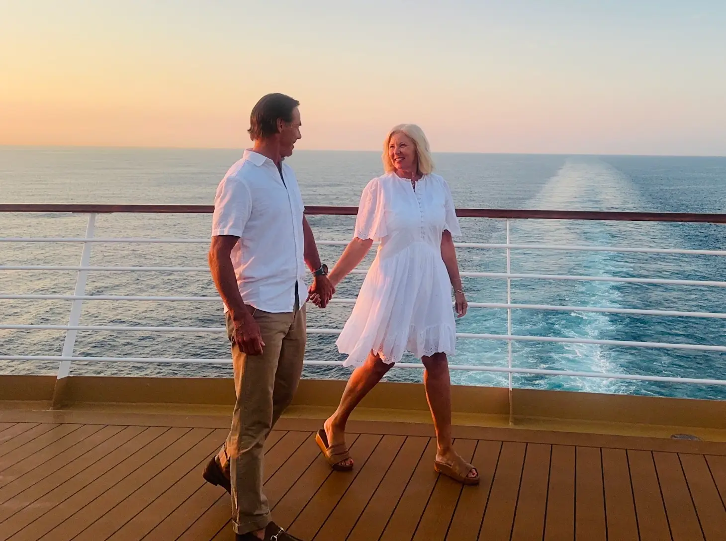The writer and her husband, dressed in white, walking across a cruise deck in front of a body of water.