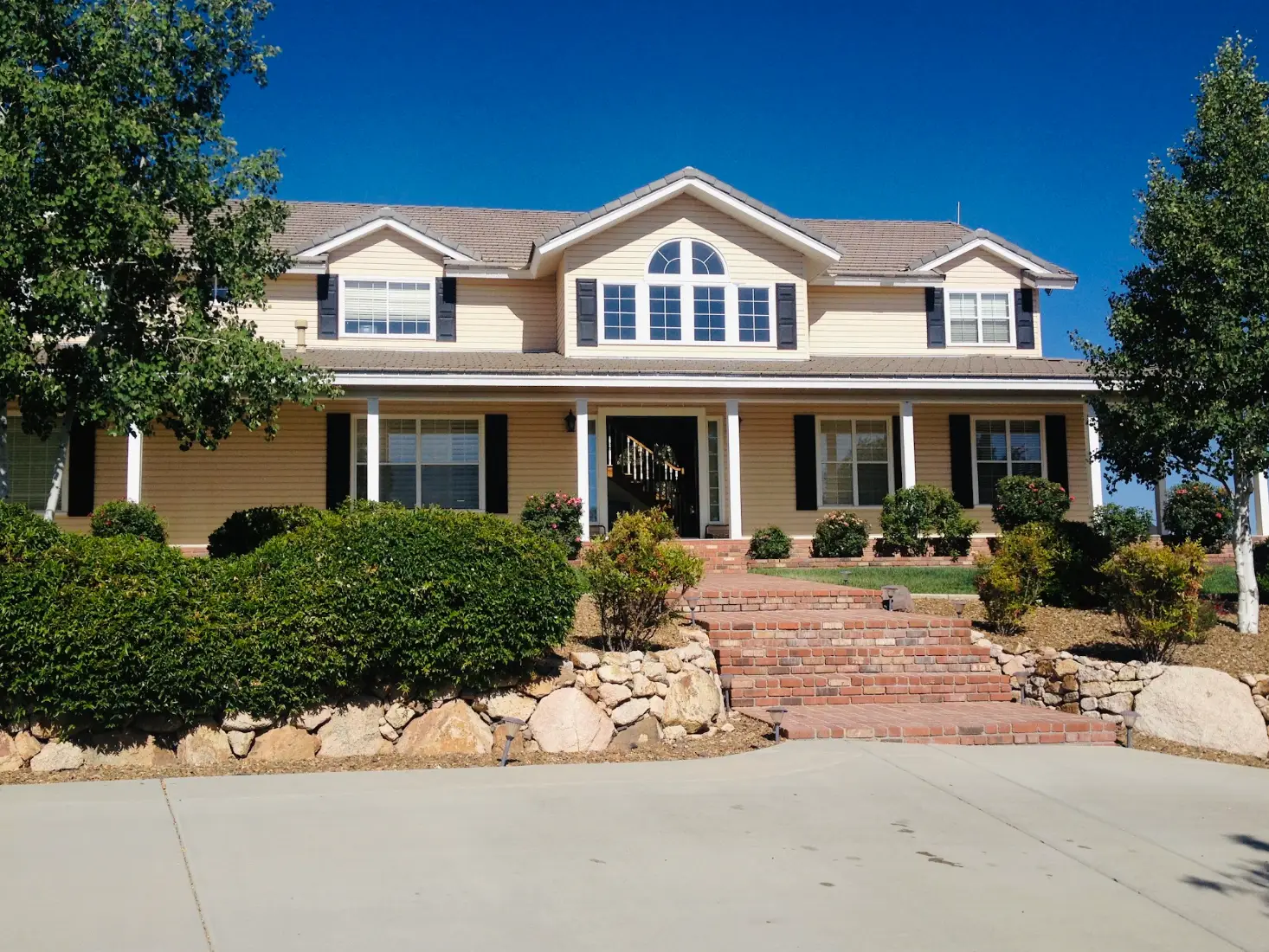 Two-story suburban house with black shutters, landscaped shrubs, brick steps, and a wide driveway under a clear blue sky.