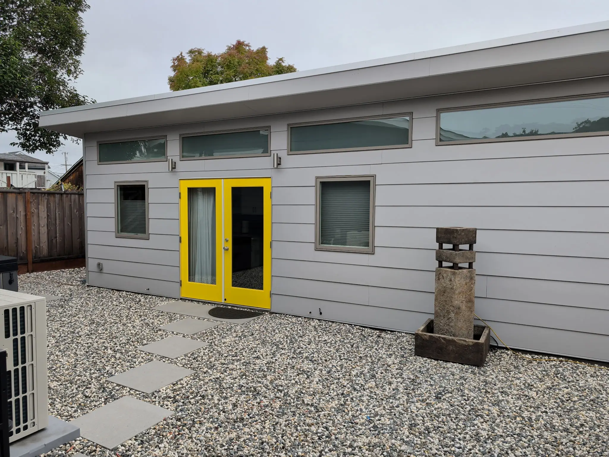 Gray modern building exterior with bright yellow double doors, narrow windows, a gravel yard, stepping stones, and a fence.