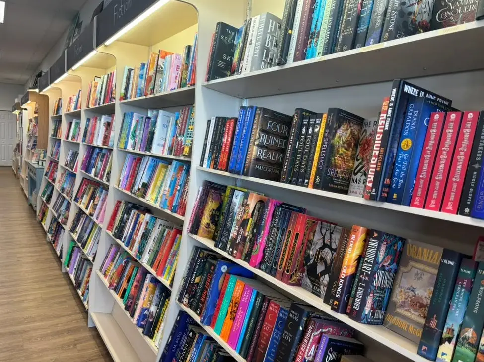 Rows of brightly lit bookshelves display colorful fiction titles in a bookstore aisle.