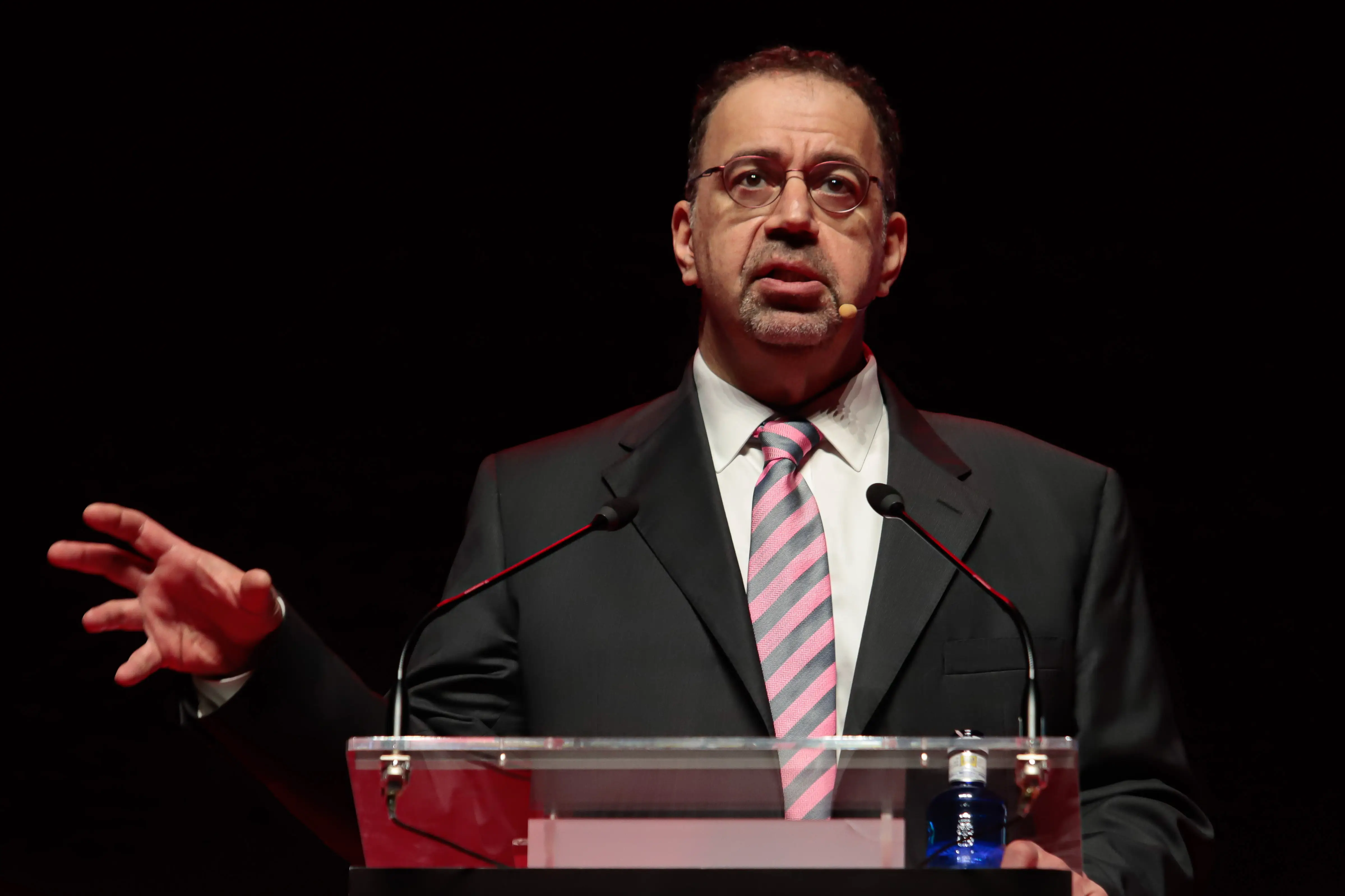A suited speaker with glasses and a striped tie gestures at a clear podium on a dark stage.