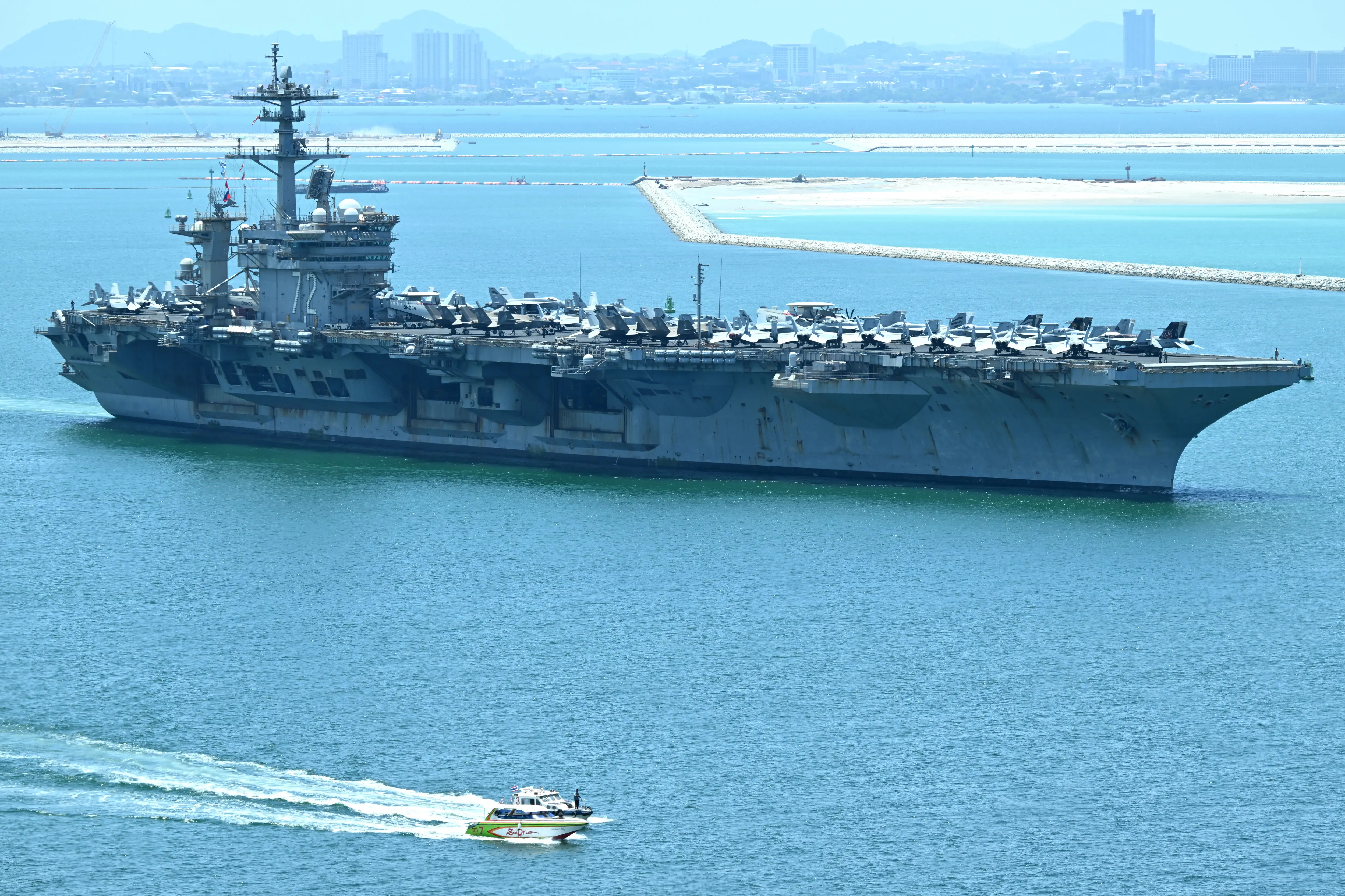 Aircraft carrier marked 76 carries rows of aircraft while a small boat speeds across blue harbor water.