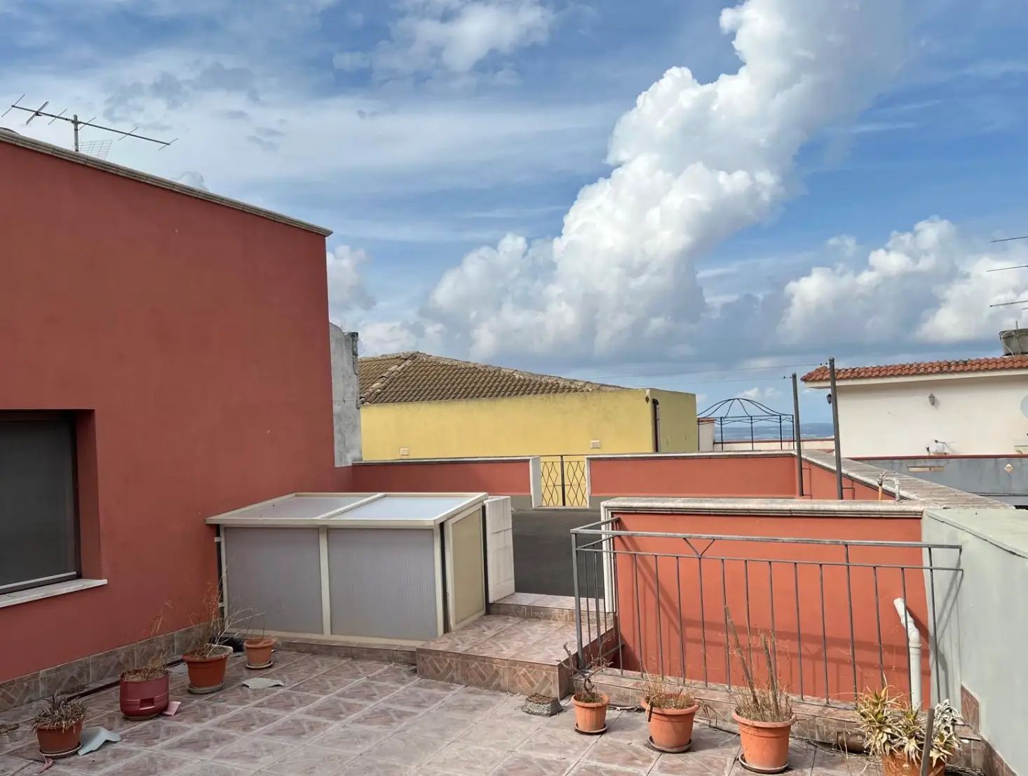Rooftop terrace with potted plants, metal railing, colorful buildings, and large clouds under a blue sky.