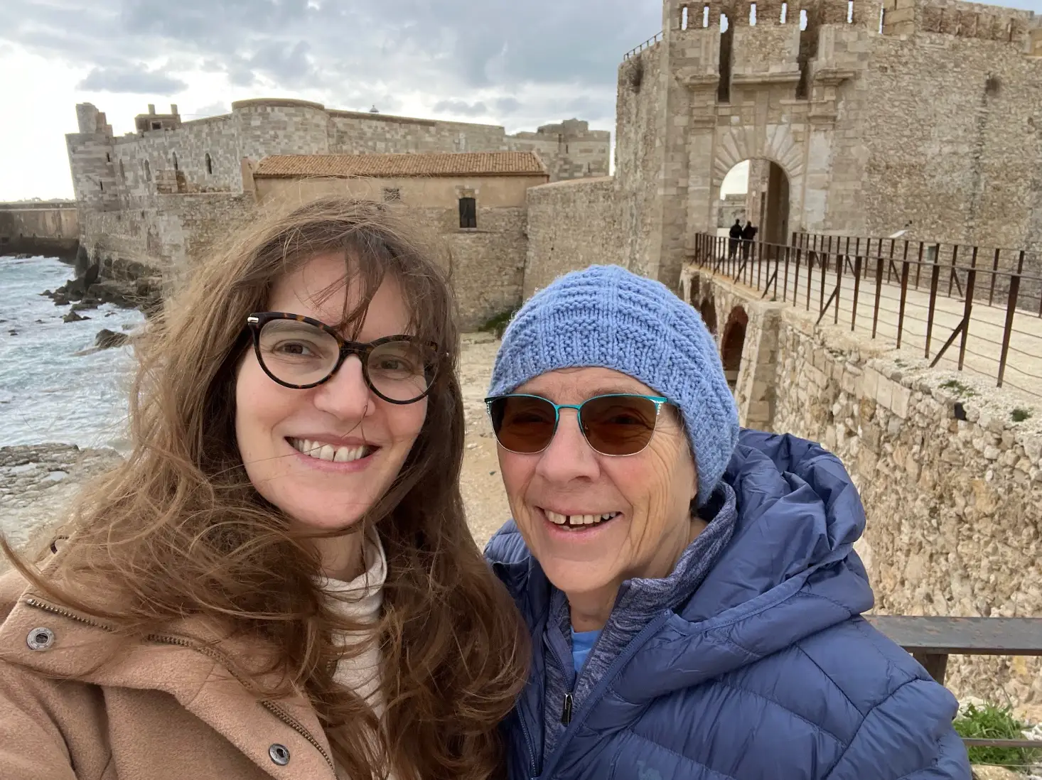 The writer and her mom take a selfie beside stone coastal fortifications and an arched gate under cloudy skies.
