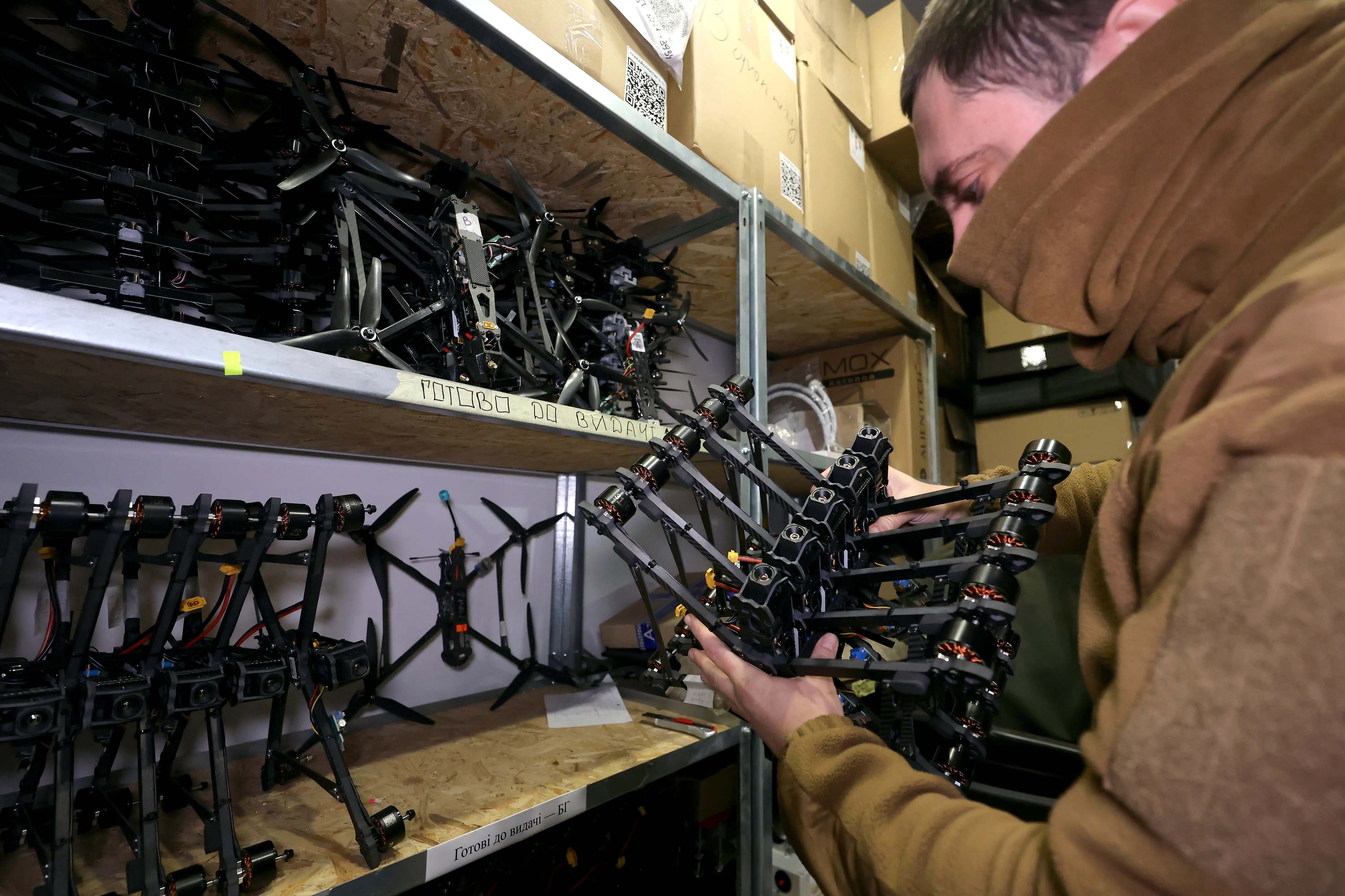 Person assembles a black multirotor drone frame on workshop shelves filled with drone parts and cardboard boxes.