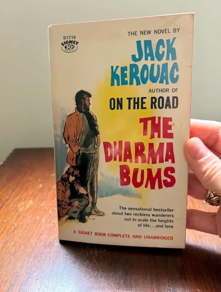 A hand holds a vintage Signet paperback of Jack Kerouac’s The Dharma Bums on a wooden surface.