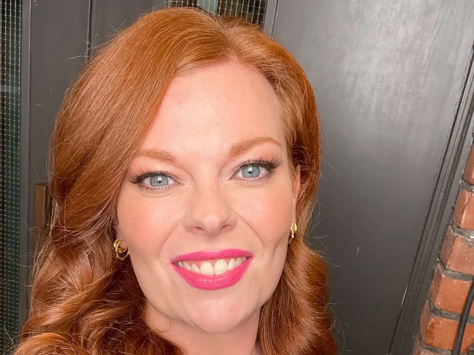 A smiling person with red hair takes a close-up selfie near a brick wall and dark door.