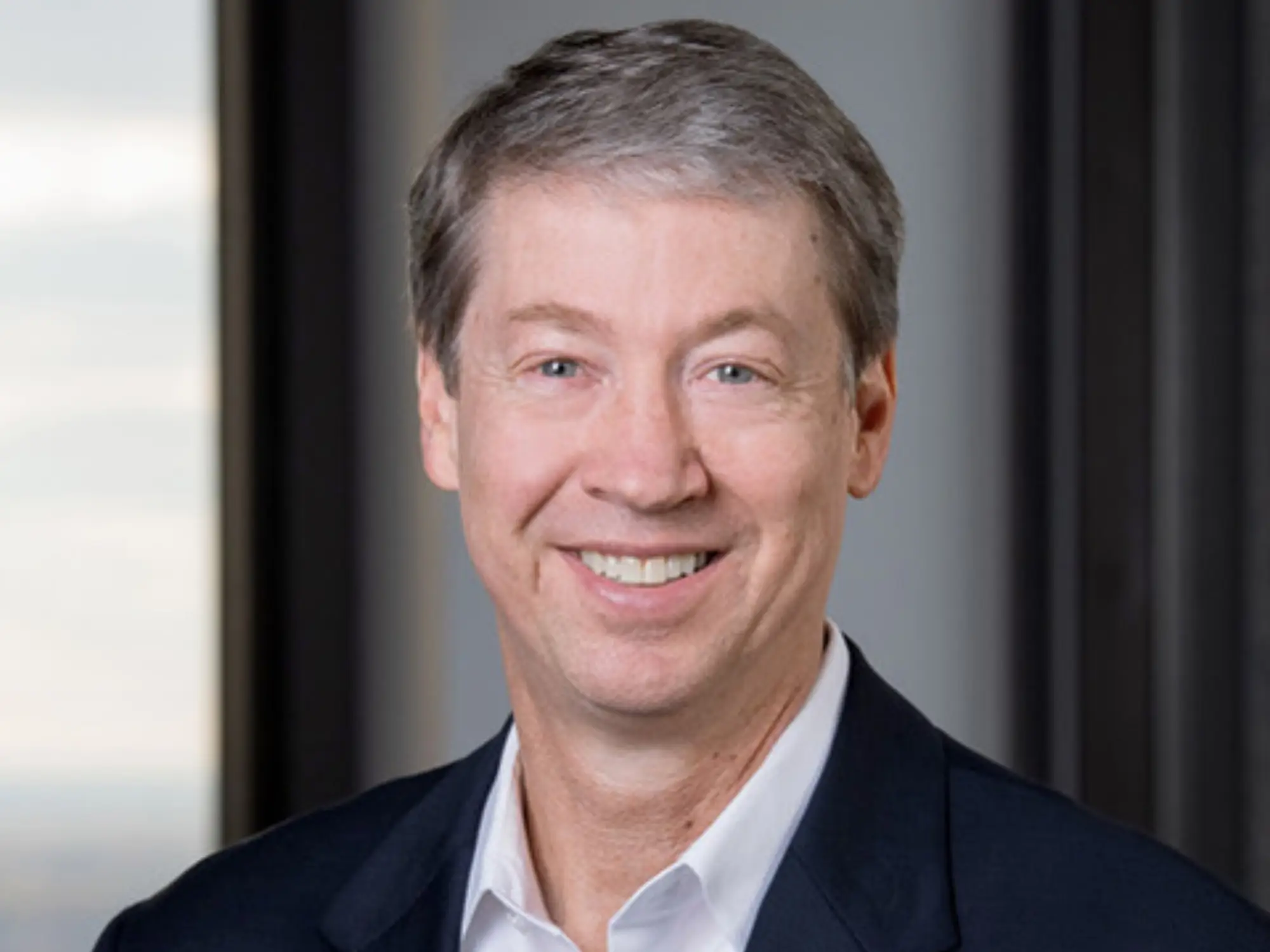 Smiling person in a dark suit jacket and white shirt poses for a professional headshot indoors.