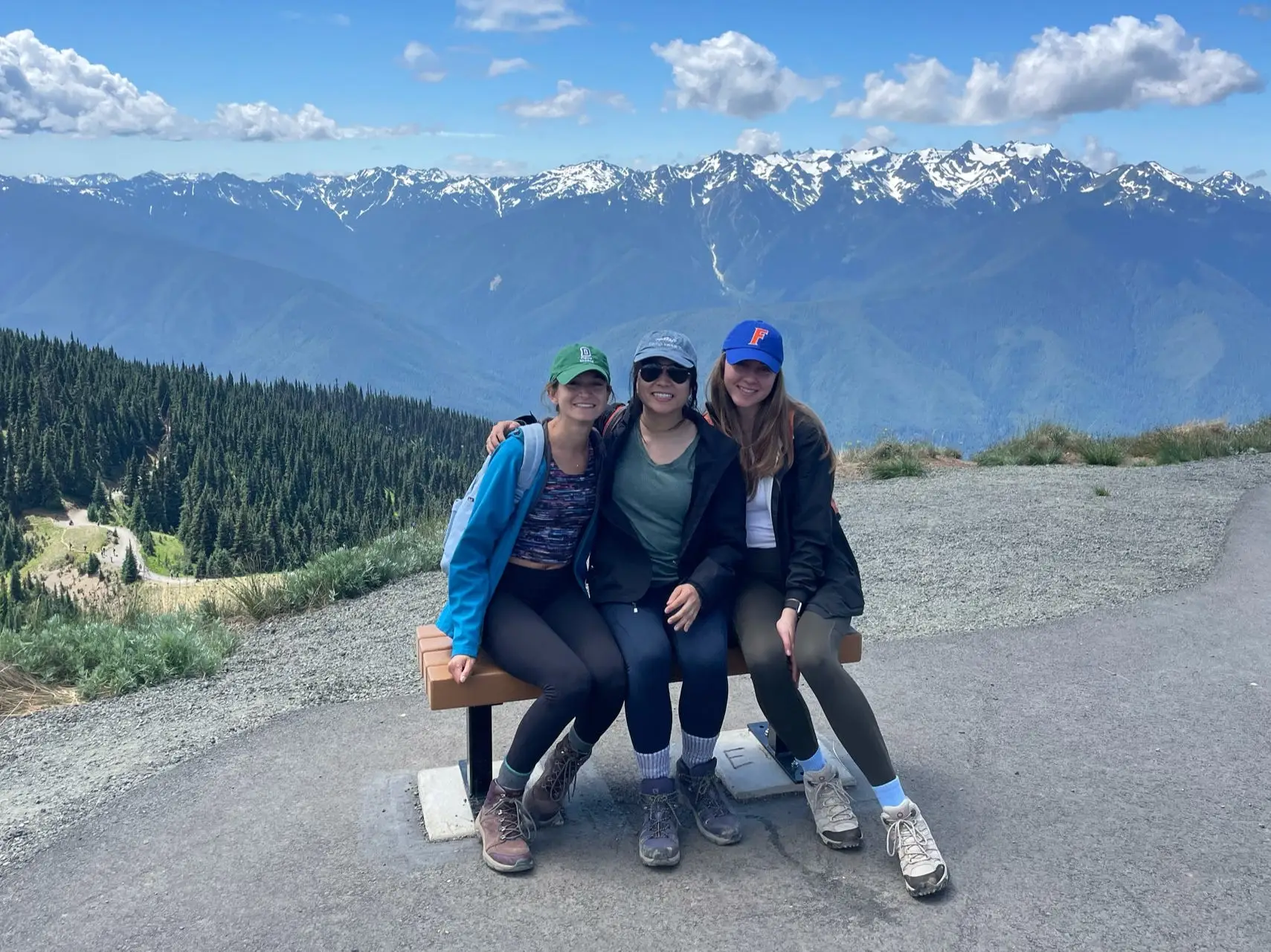 Anna and two friends sit on a bench in Olympic National Park. Behind them are mountains and trees.
