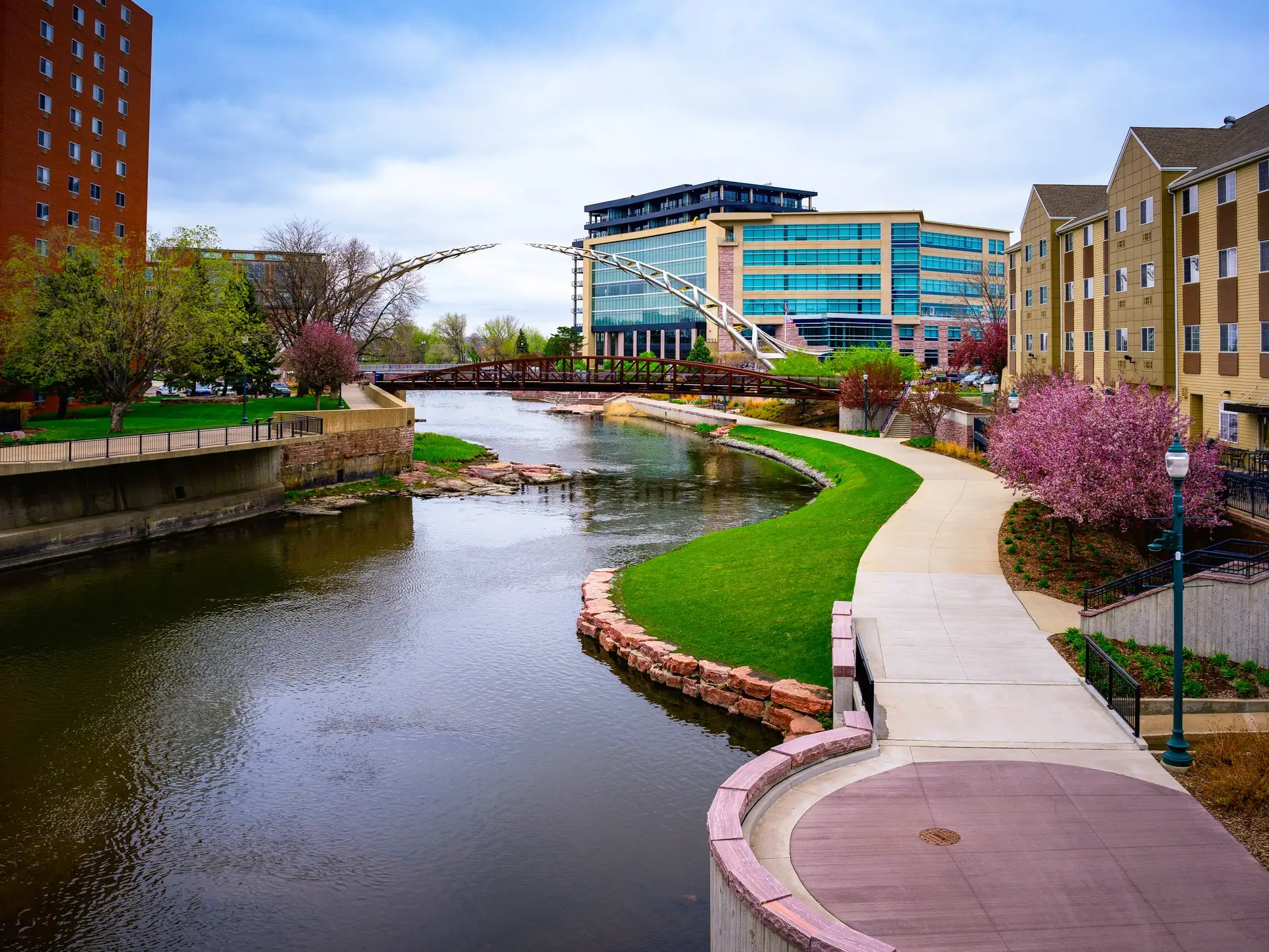 Riverwalk curves beside a downtown river with bridges, spring trees, and the Arc of Dreams sculpture.