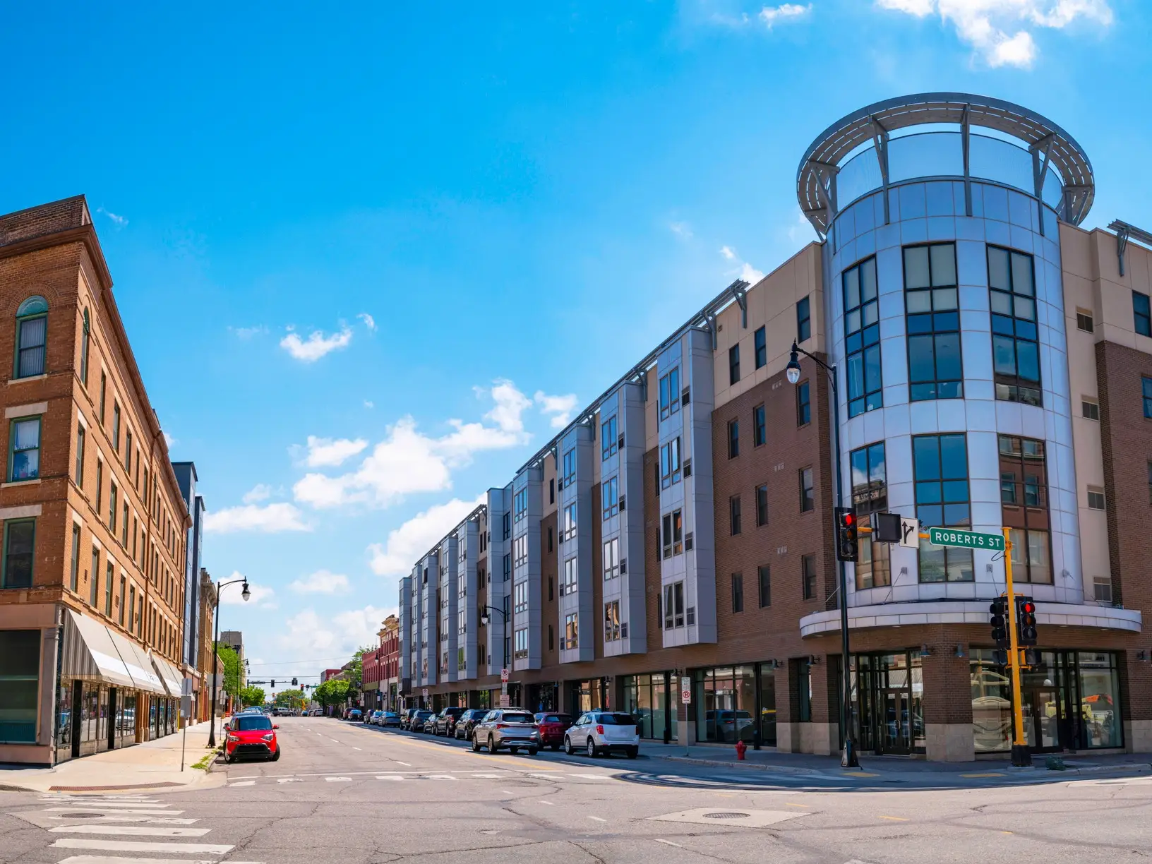 Modern apartment buildings and storefronts line a city street at an intersection marked ROBERTS ST under a blue sky.