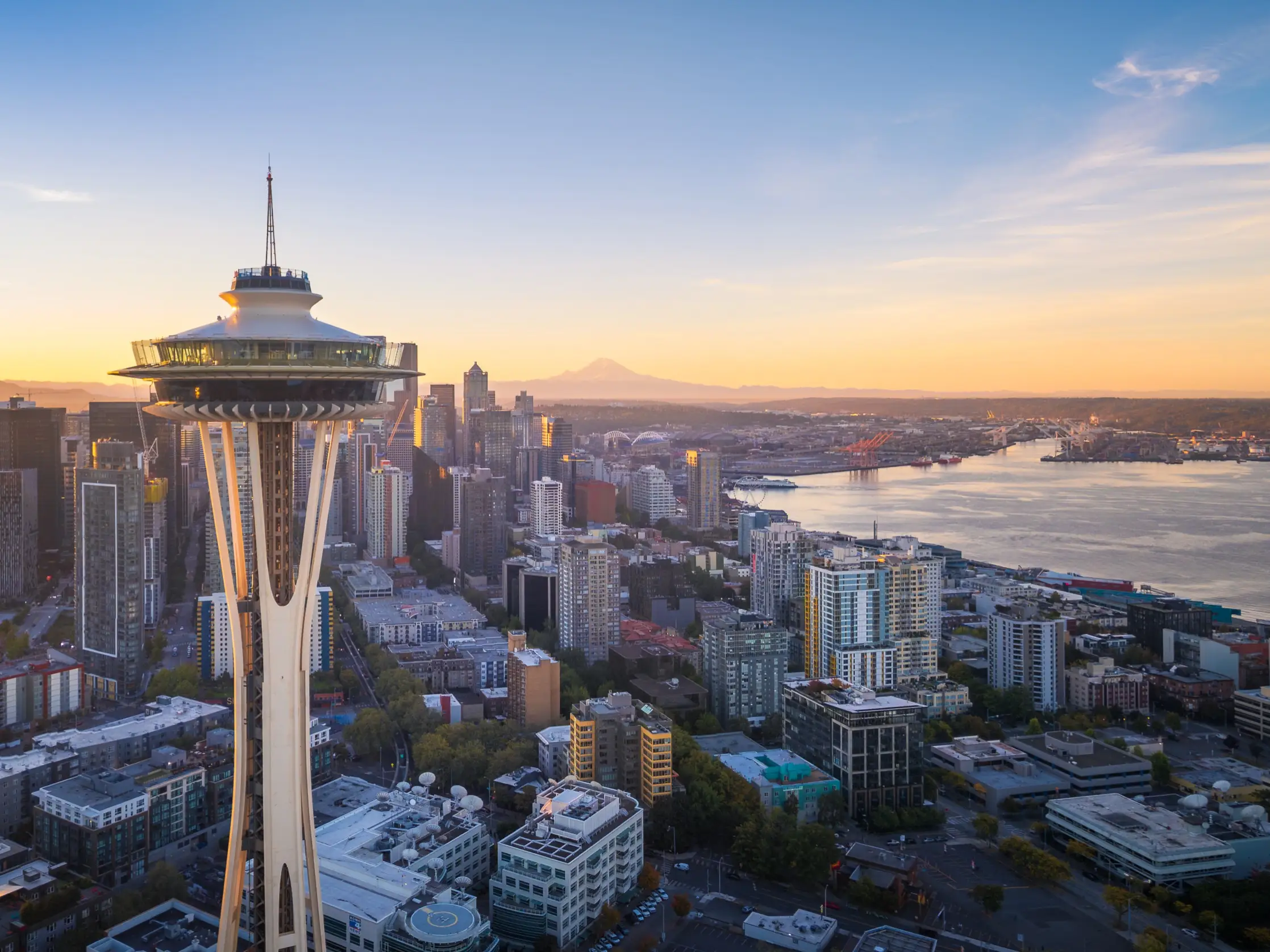 Seattle skyline at sunset with the Space Needle, Elliott Bay waterfront, and Mount Rainier in the distance.