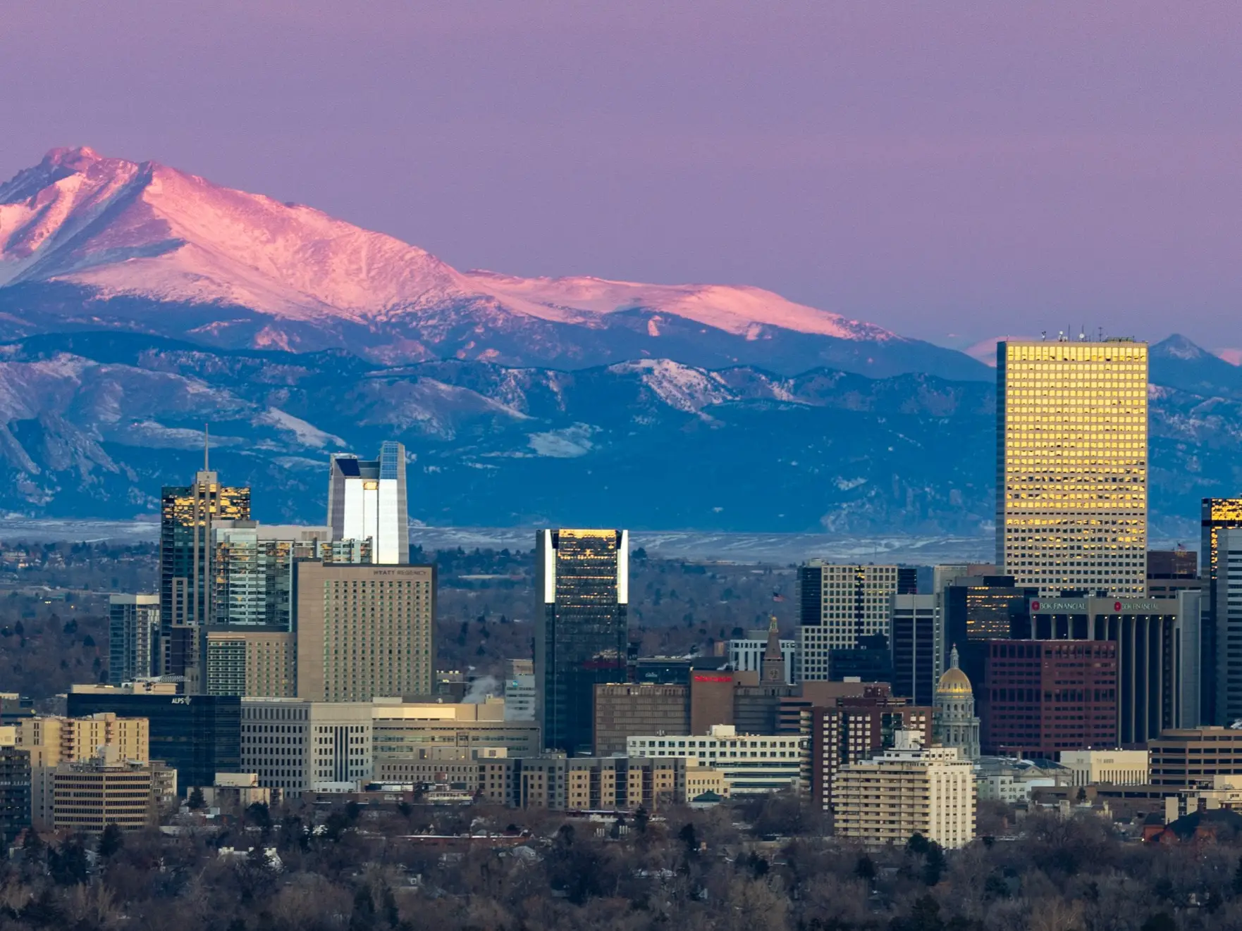 Downtown Denver skyline stands before snow-covered Rocky Mountains under pink sunset light.