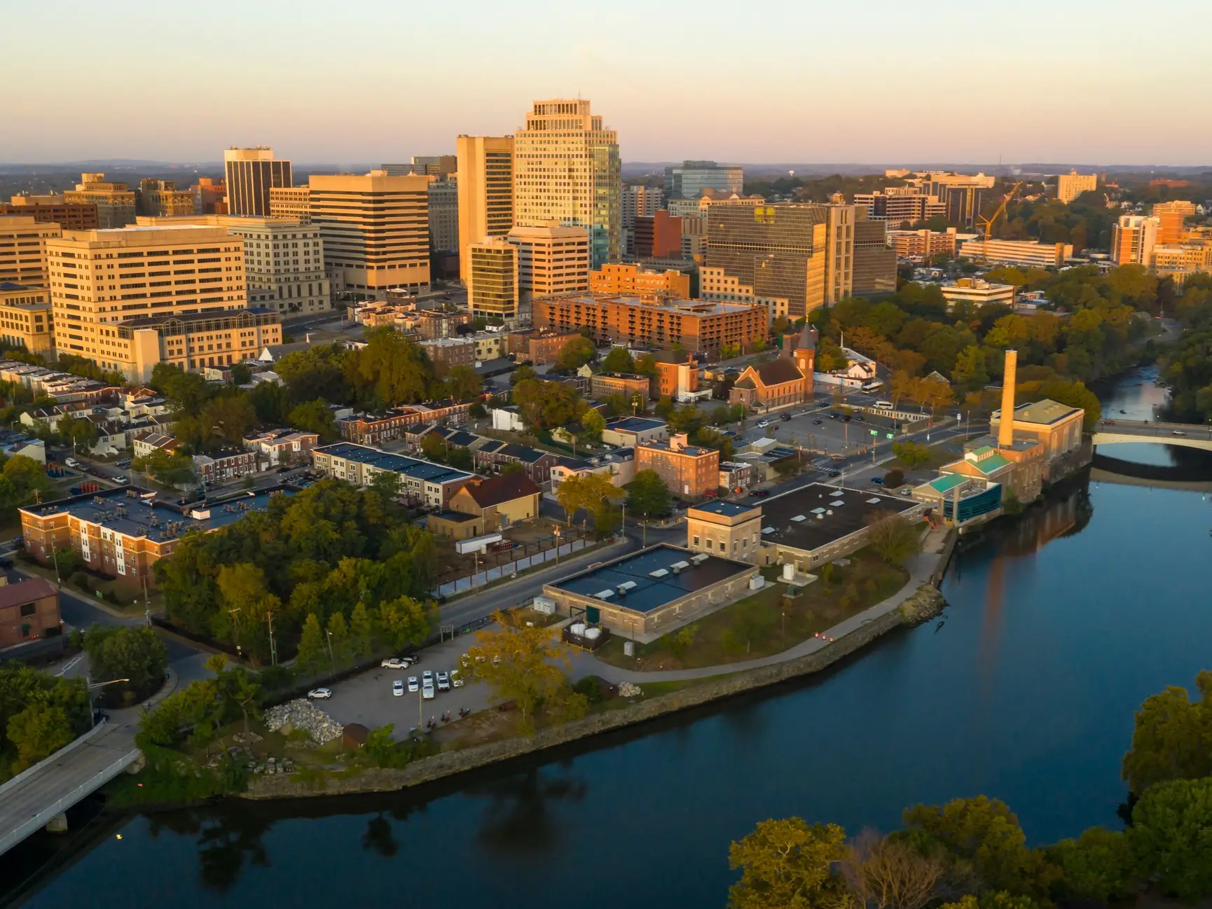 Downtown skyline and waterfront buildings are lit by warm sunset light beside a winding river.