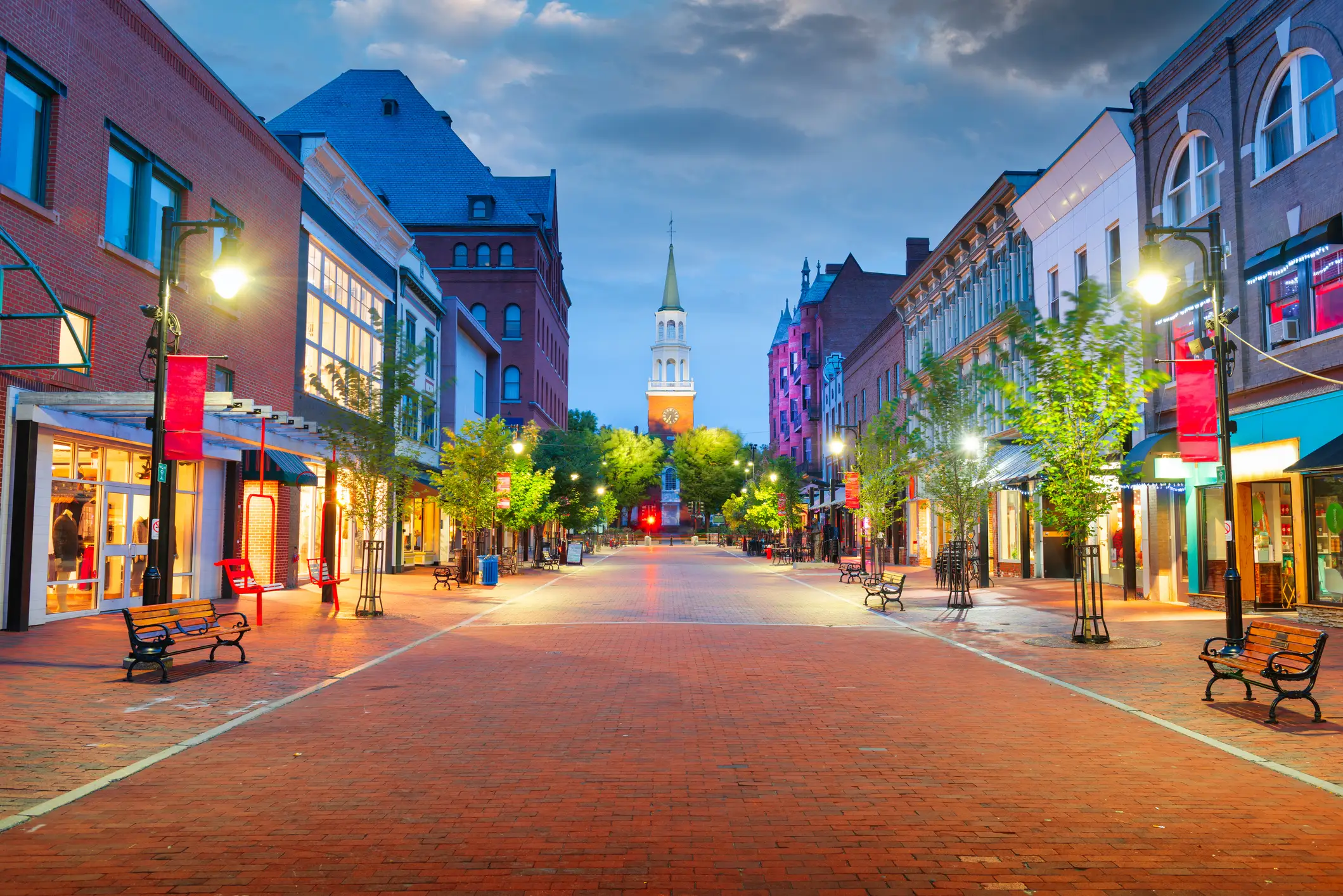 Brick pedestrian shopping street at dusk with lit storefronts, benches, trees, and a white church steeple in the distance.