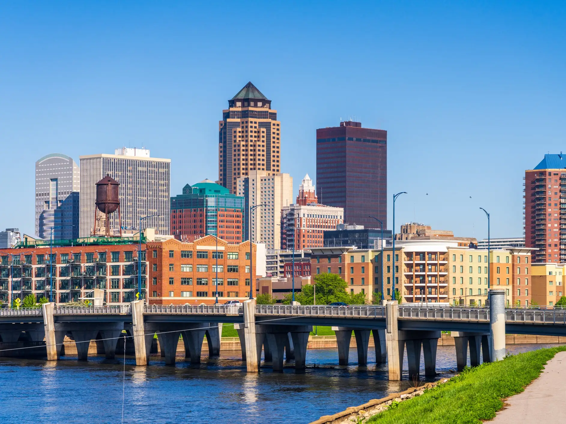 Downtown Des Moines skyline rises behind a river bridge under a clear blue sky.