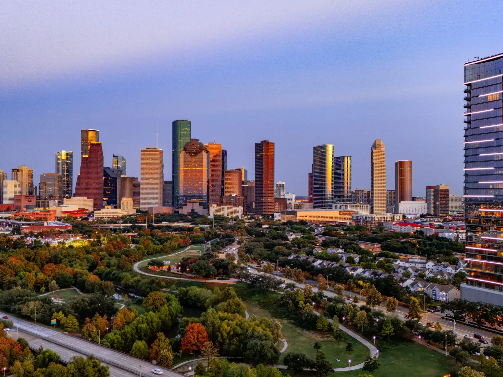Downtown Houston skyline rises above parks, roads, and residential buildings at dusk.