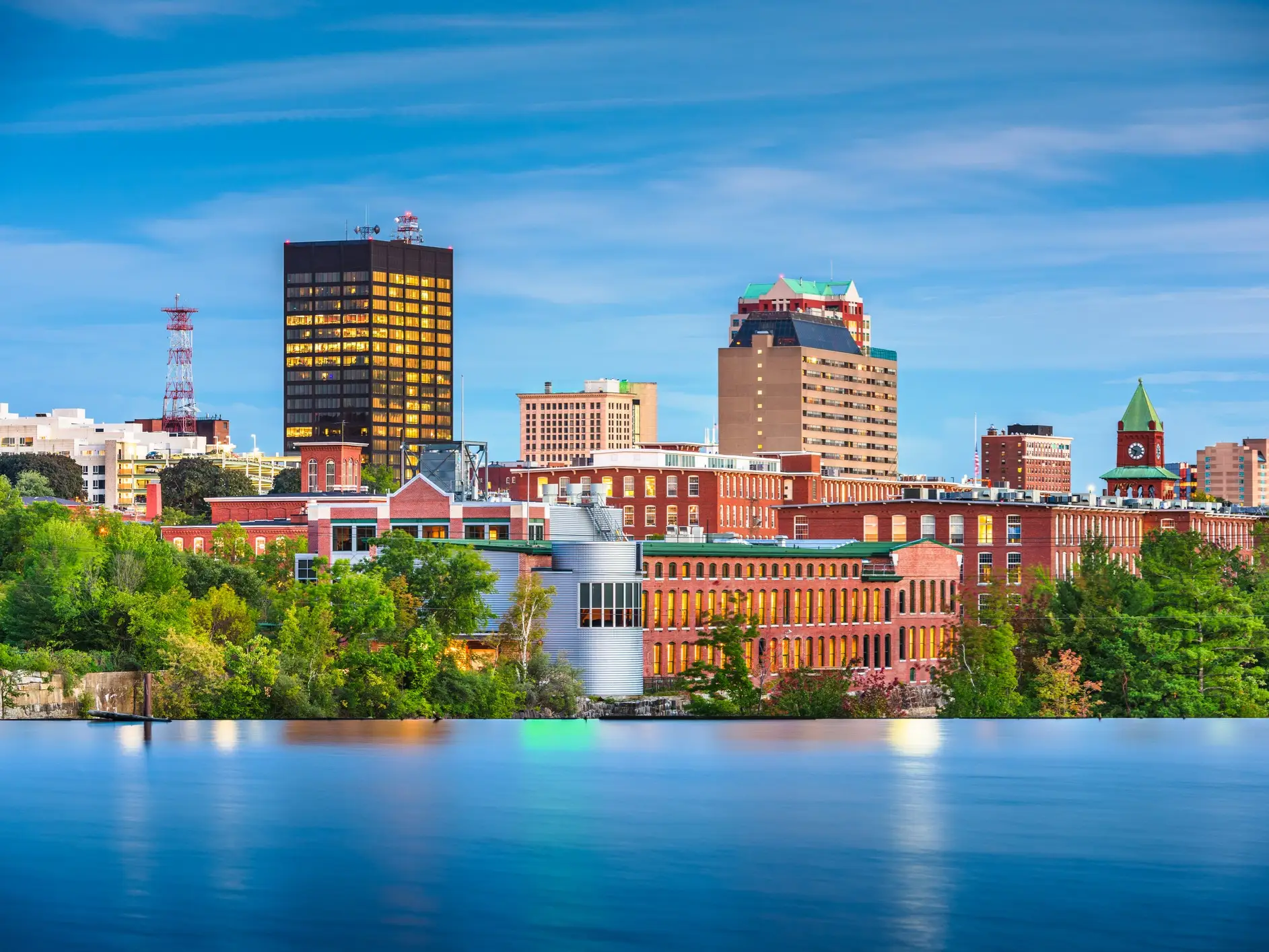 Manchester, New Hampshire skyline and red brick mill buildings reflect across a calm river at dusk.