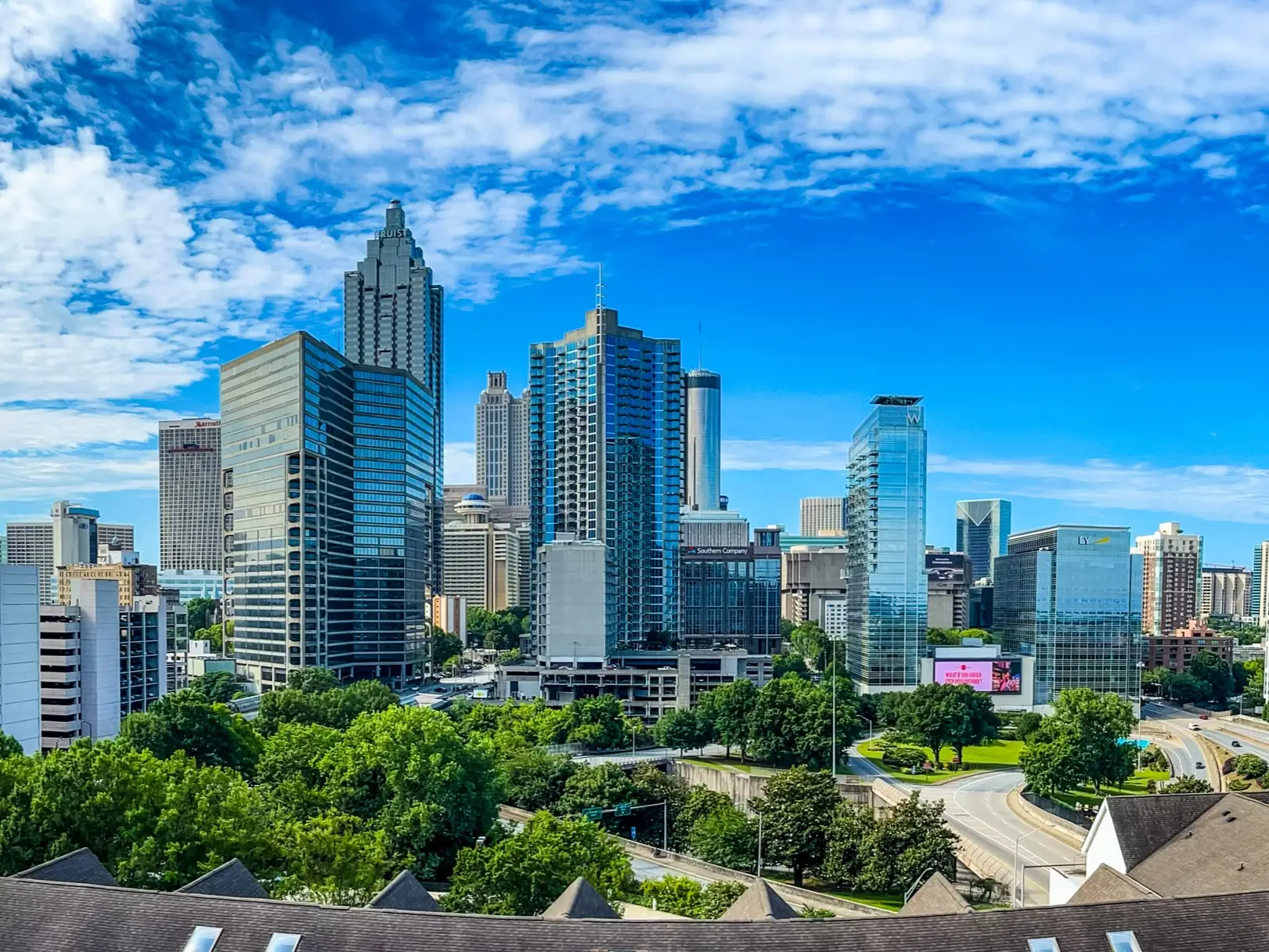 Atlanta skyline with downtown high-rises, green trees, roads, and a bright blue clouded sky.