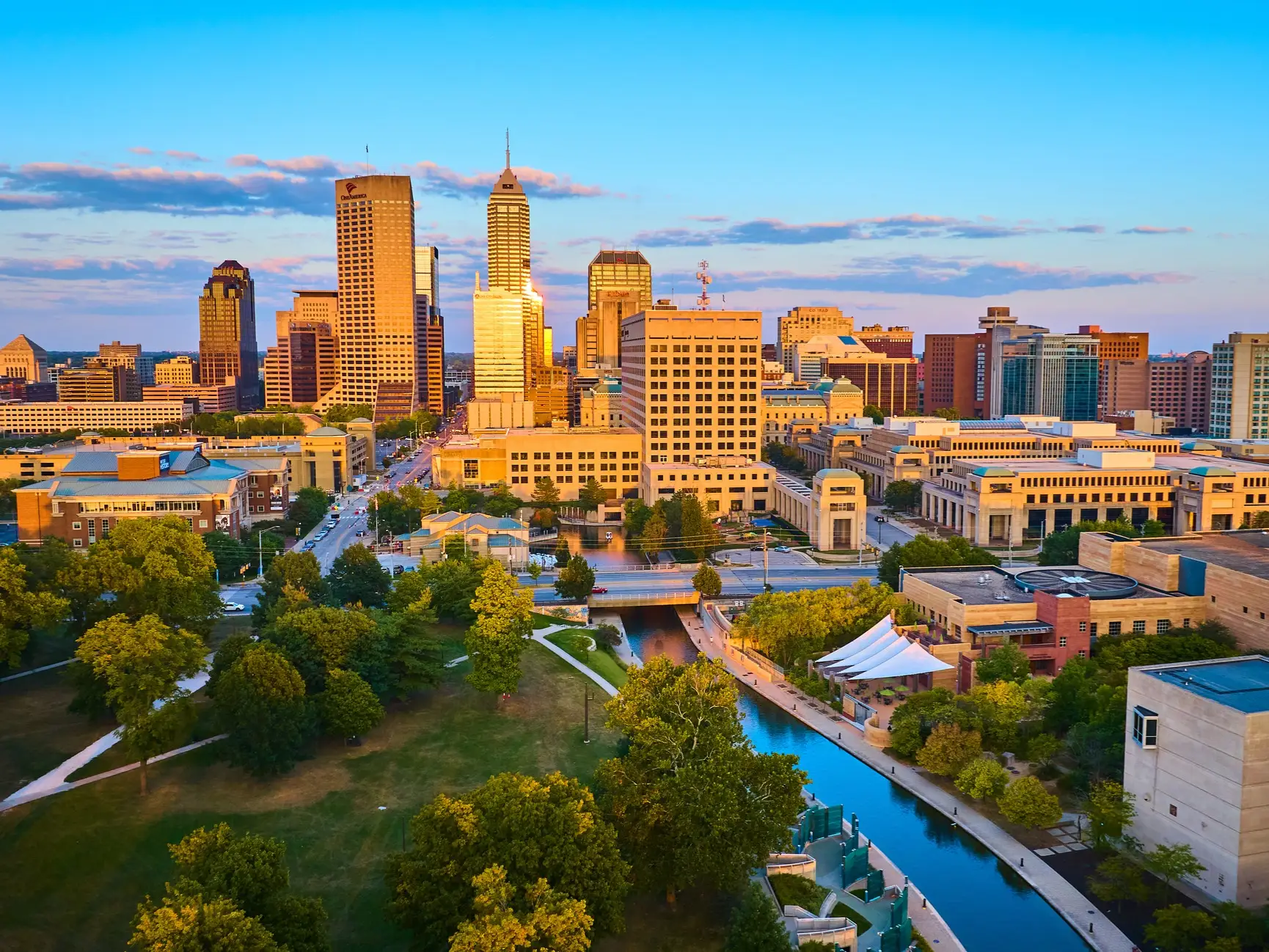 Downtown Indianapolis skyline rises above green parks and a canal in warm sunset light.
