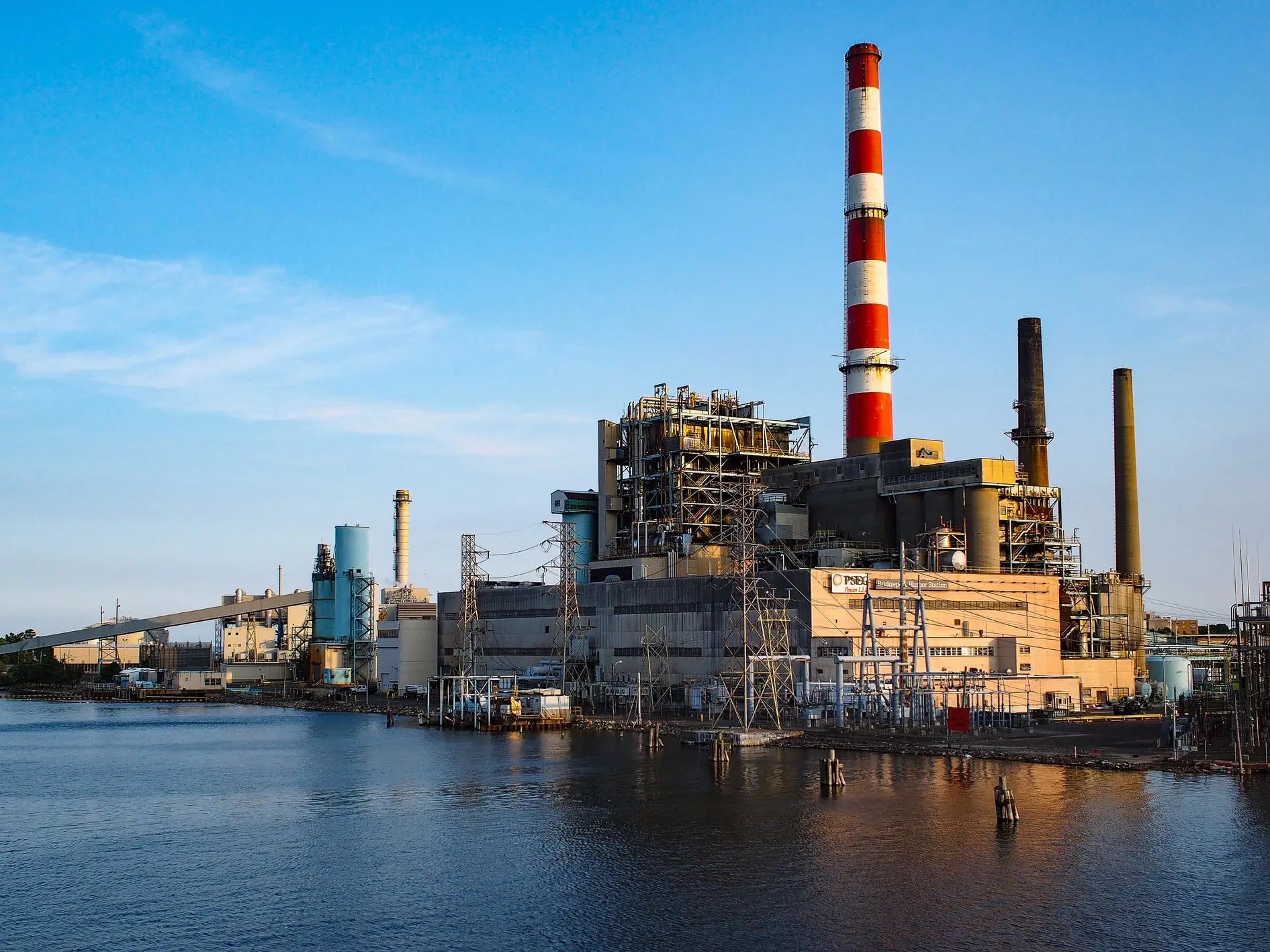 Waterfront industrial power plant with tall smokestacks and electrical equipment under a clear blue sky.