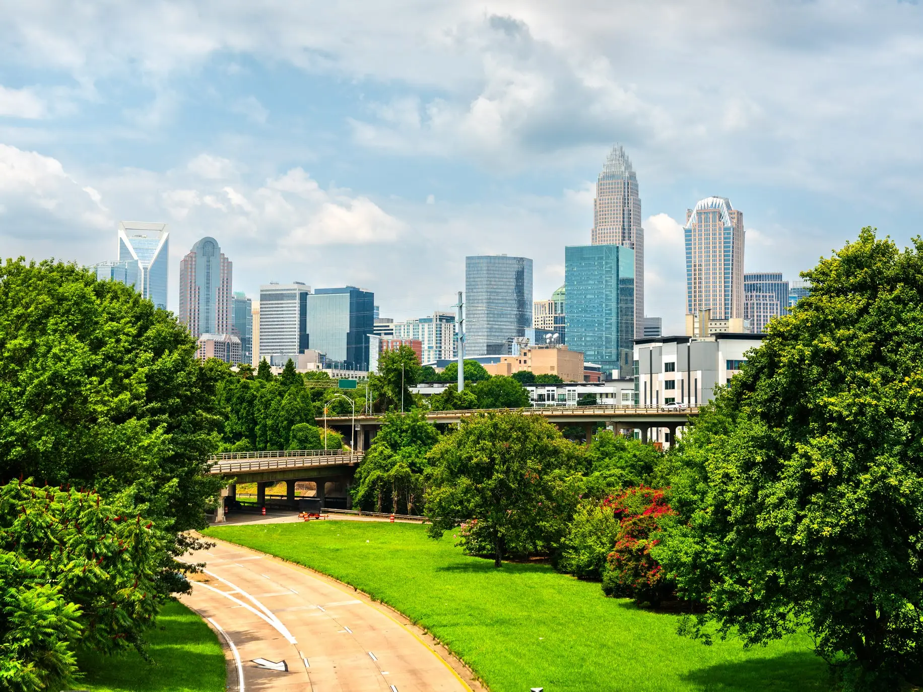Uptown Charlotte skyline rises beyond green trees, parkland and a curving roadway under a partly cloudy sky.