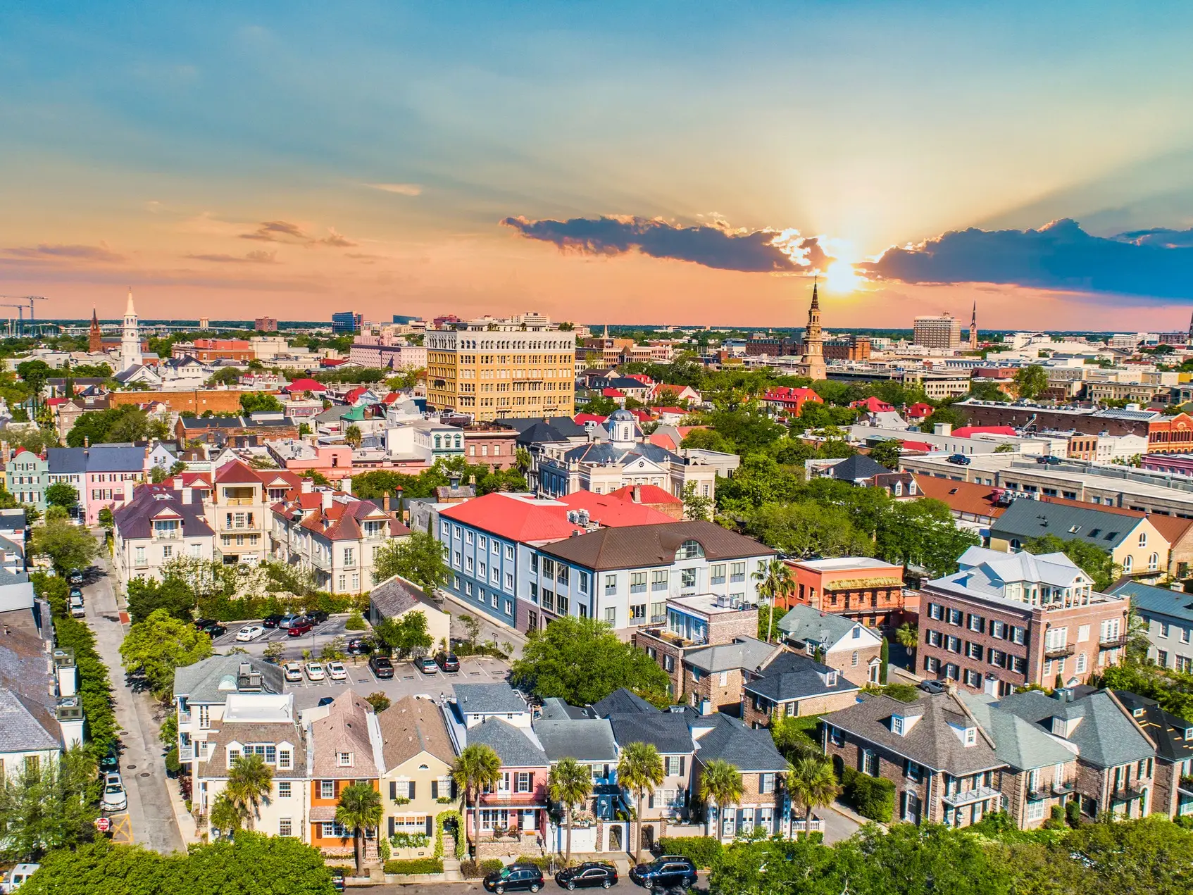 Aerial view of historic Charleston rooftops, church steeples, palm-lined streets, and downtown buildings at sunset.