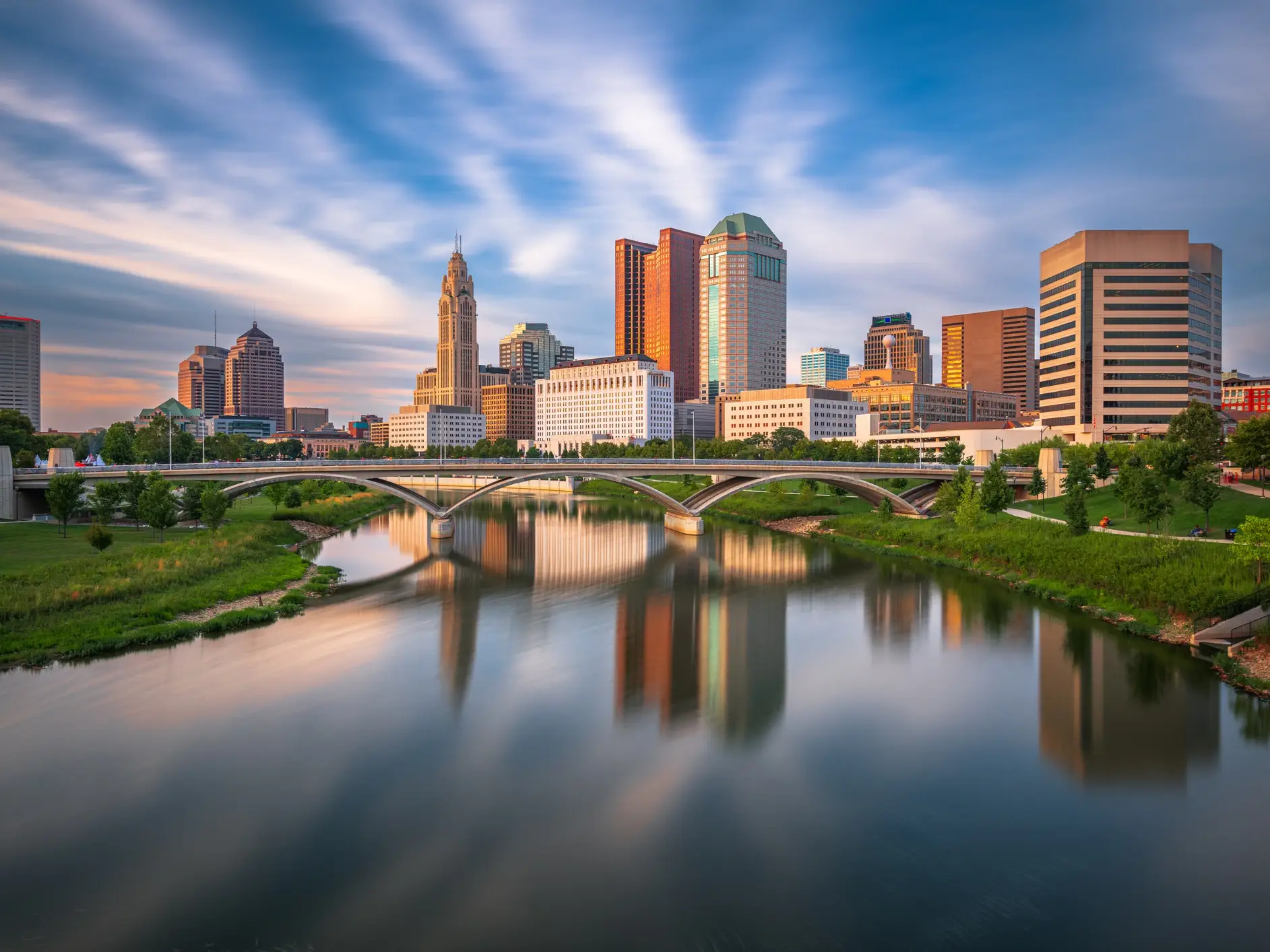Columbus skyline reflects in the Scioto River at sunset with an arched bridge in the foreground.