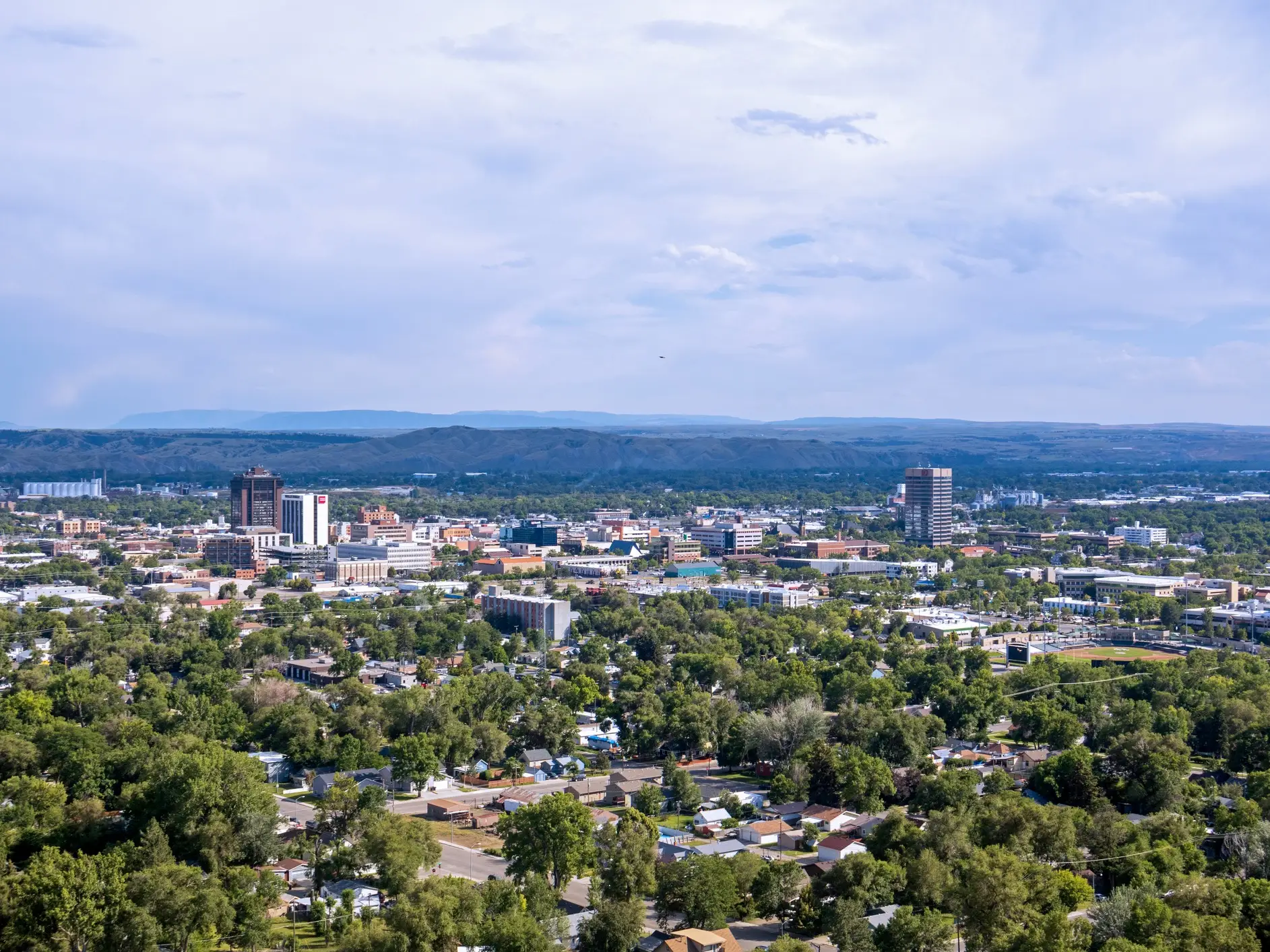 Downtown skyline and tree-filled neighborhoods stretch toward distant hills under a cloudy blue sky.