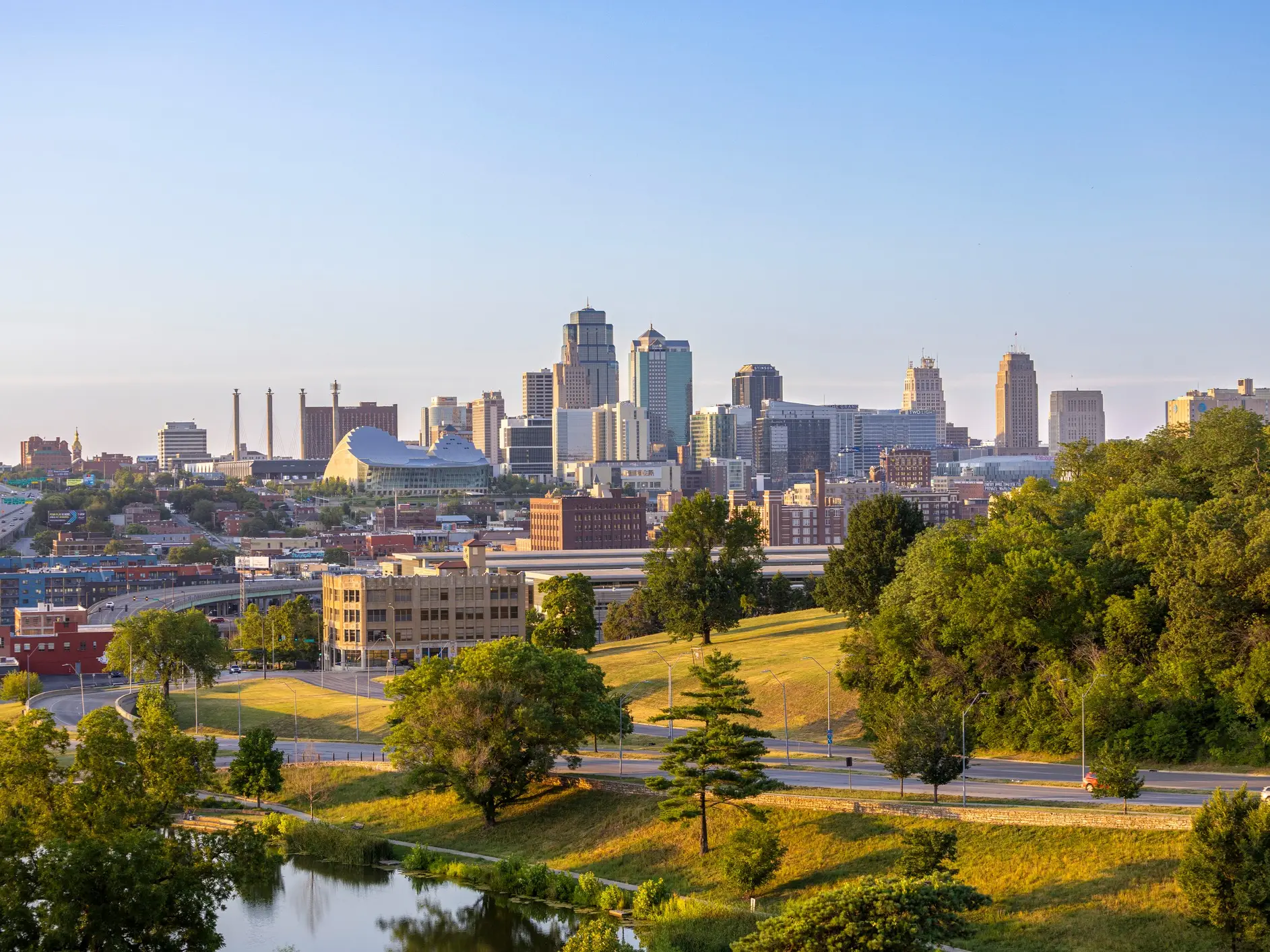 Downtown Kansas City skyline rises beyond green parkland and roads in warm evening light.