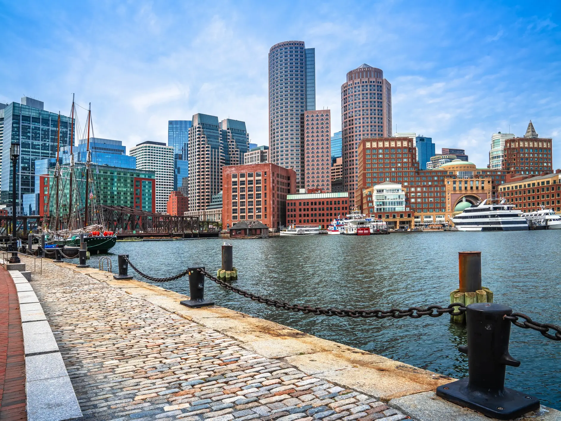 Boston waterfront skyline with boats, a cobblestone harborwalk, chain posts, and high-rise buildings along the water.